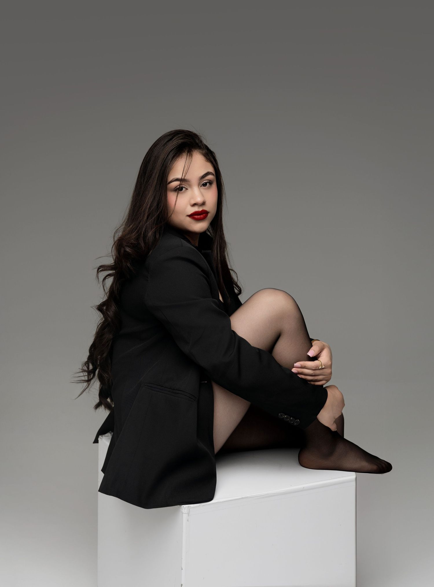Young woman in black blazer and stockings sits on white cube, posing confidently with red lips and long dark hair.