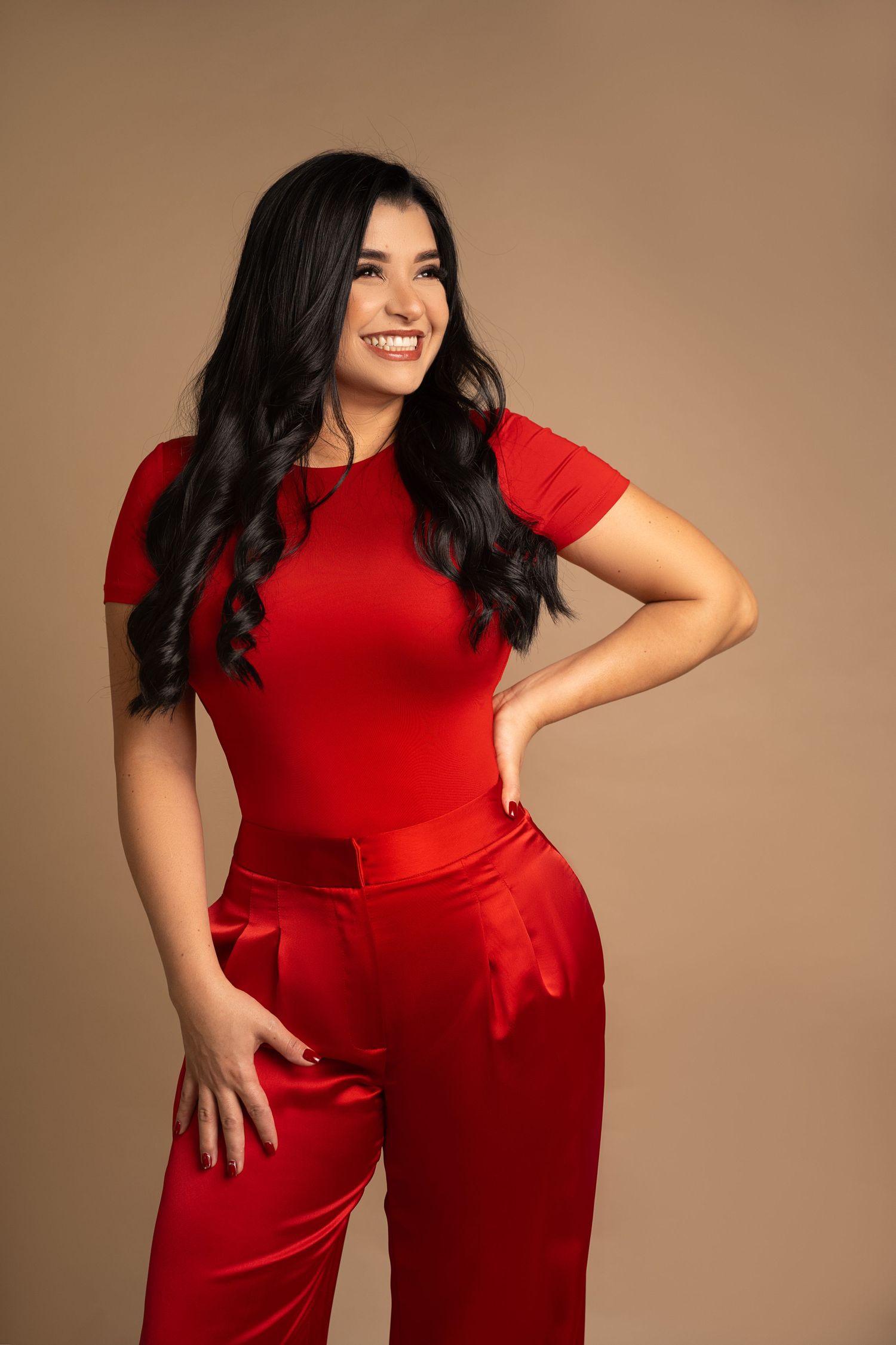 Smiling young woman in a red top and satin red pants posing confidently against a beige background.