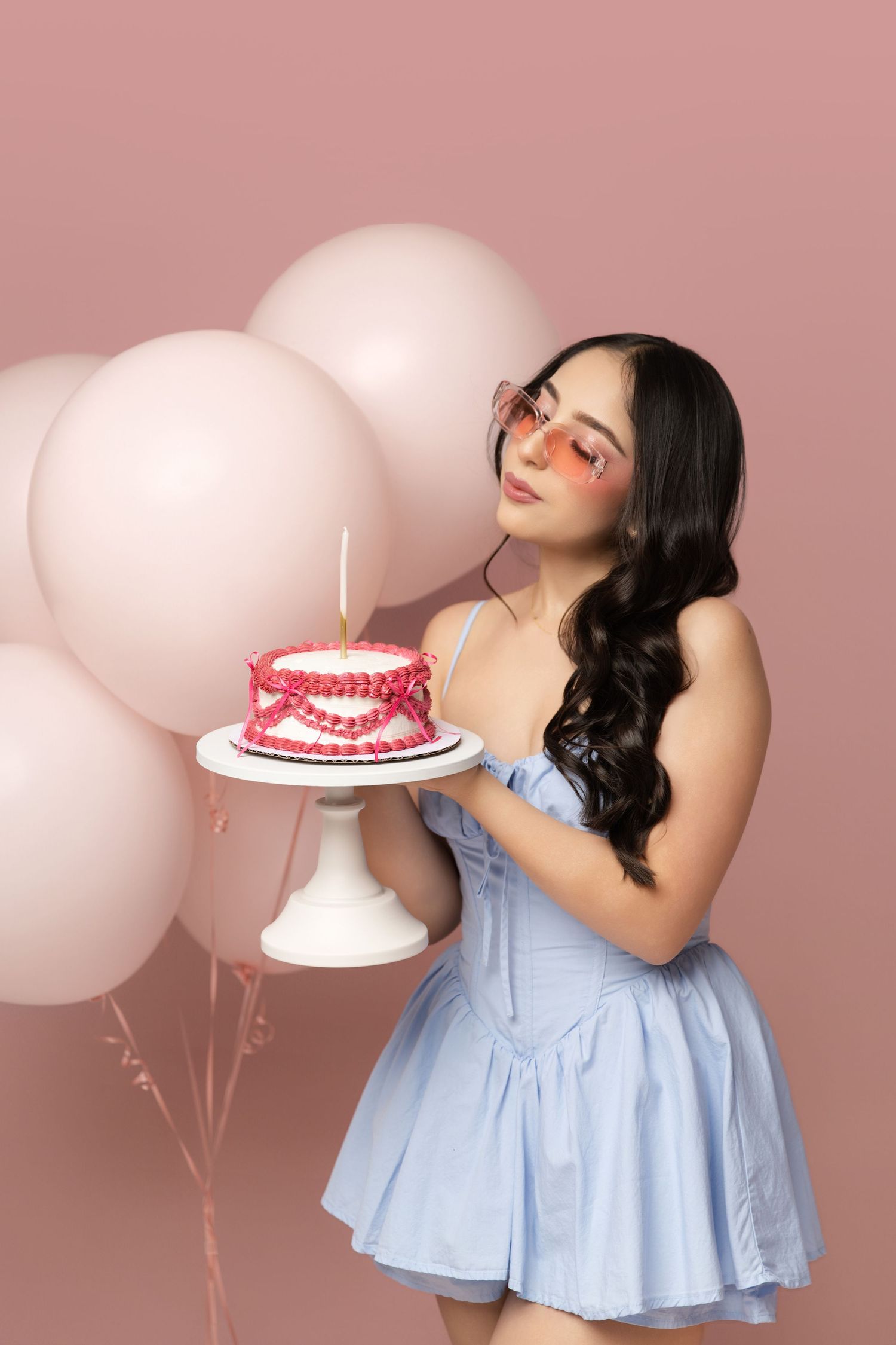 Young woman in blue dress and pink sunglasses holds birthday cake with candle, surrounded by pink balloons on pink background.