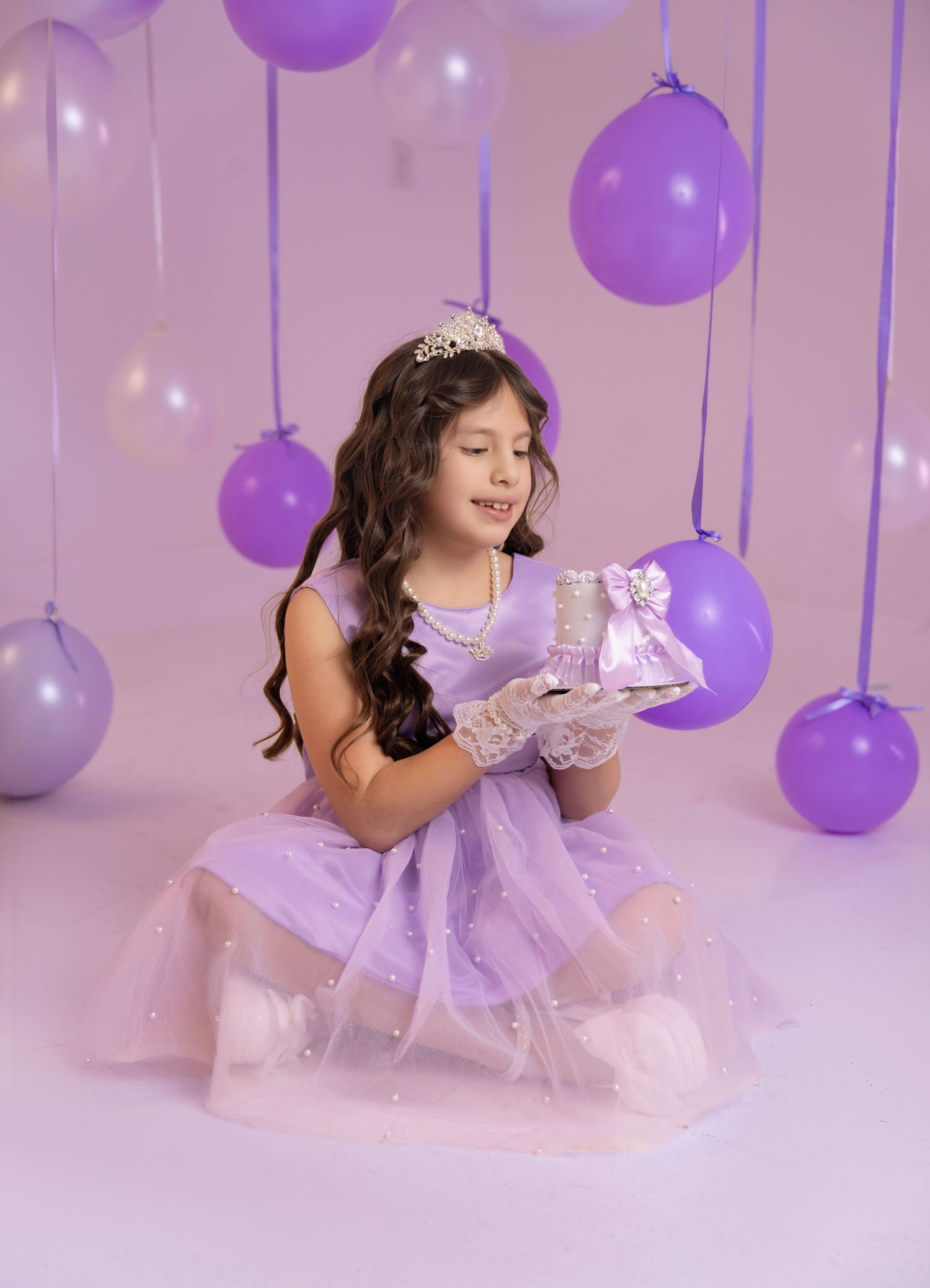 A young girl in a lavender princess dress and tiara holds a gift box, surrounded by purple balloons.