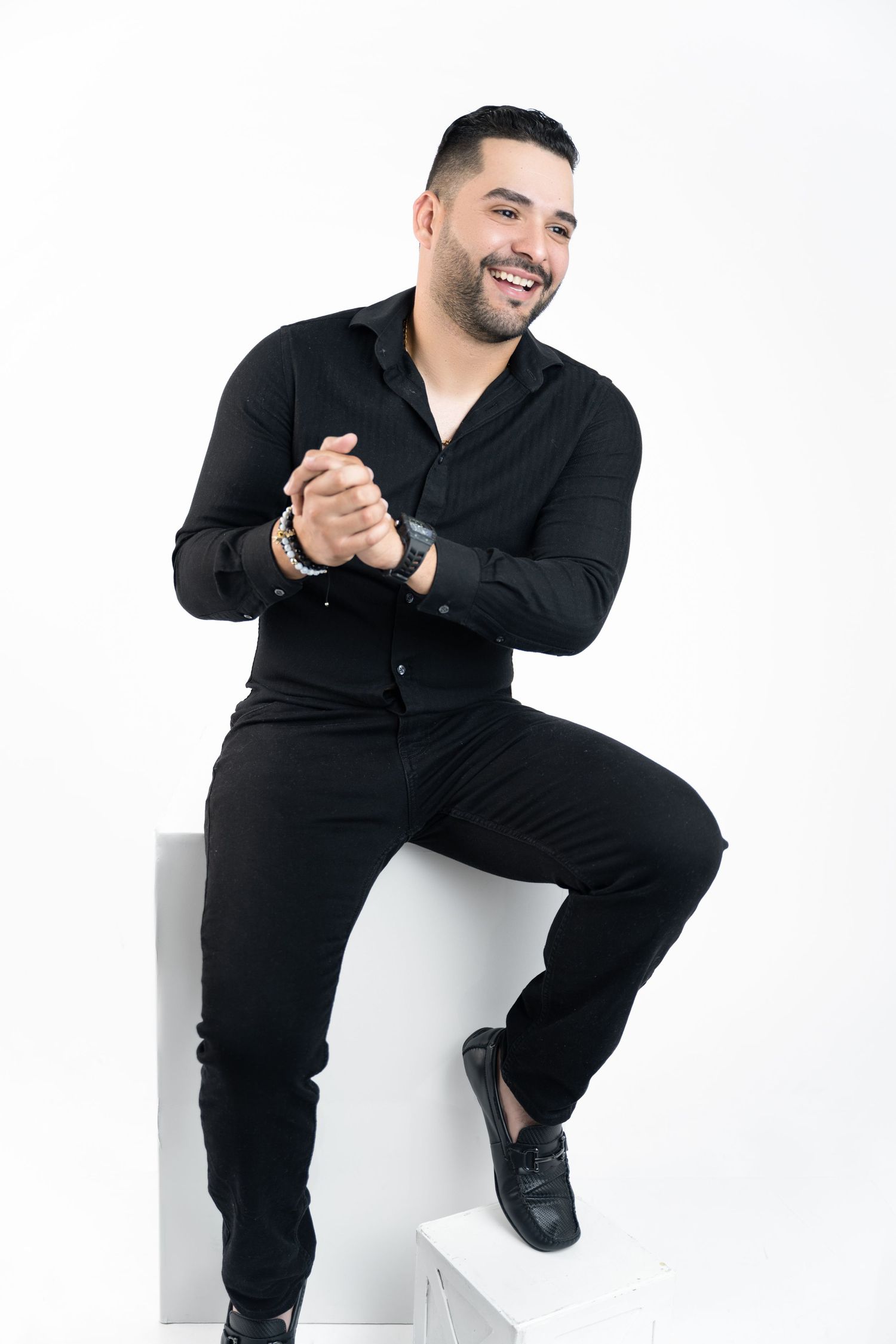 Smiling man in black shirt and pants sits on white cubes with clasped hands against a clean white studio background.