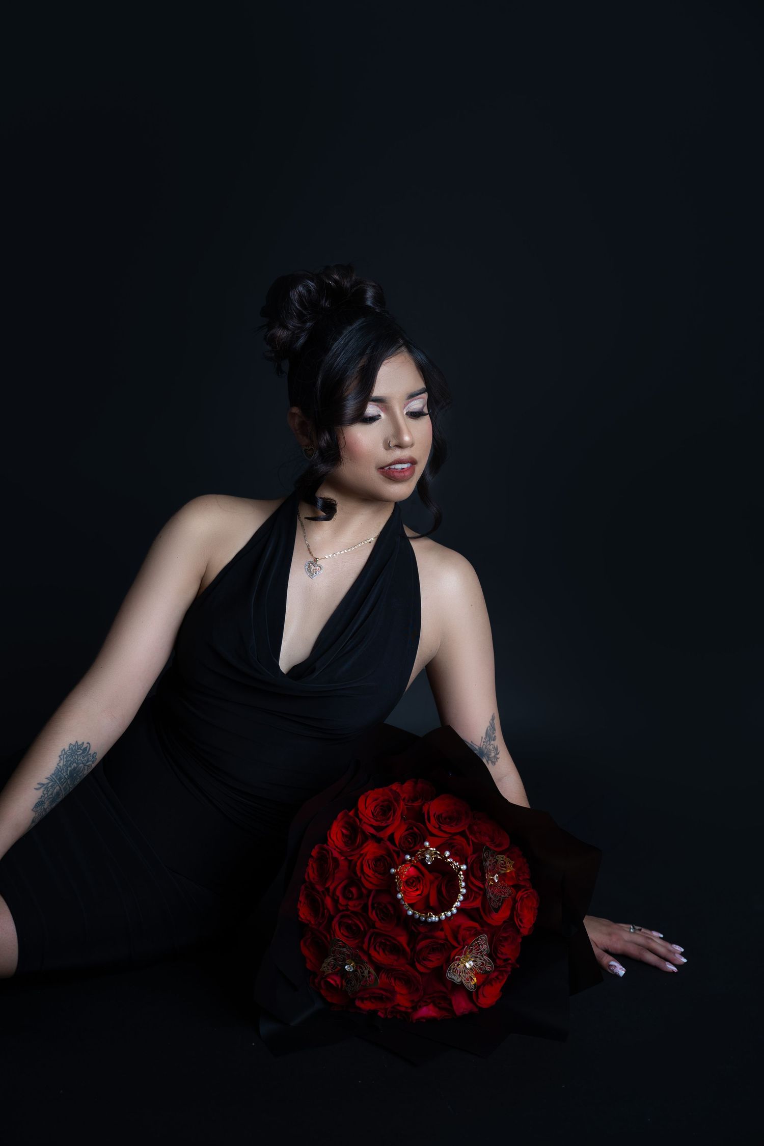 Woman in a black halter dress poses on a dark studio backdrop beside a large bouquet of red roses wrapped in black paper.