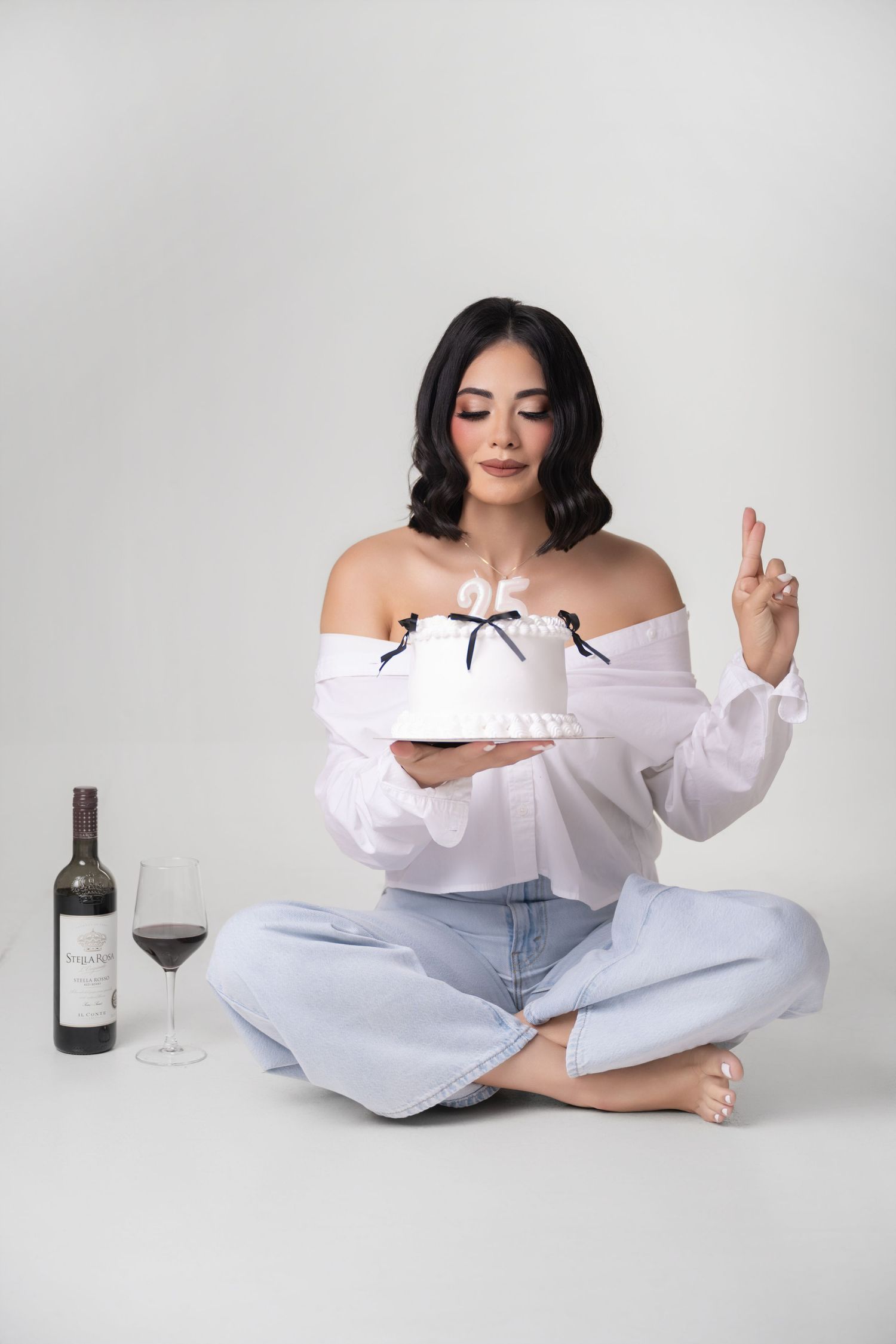 Woman in an off-shoulder white blouse and light jeans sits cross-legged holding a white bow-trimmed birthday cake beside a glass of red wine