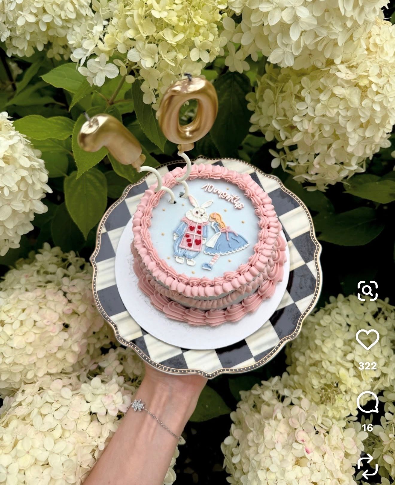 Hand holding a pink-piped birthday cake with an Alice in Wonderland rabbit design on a checkered plate before white hydrangeas.