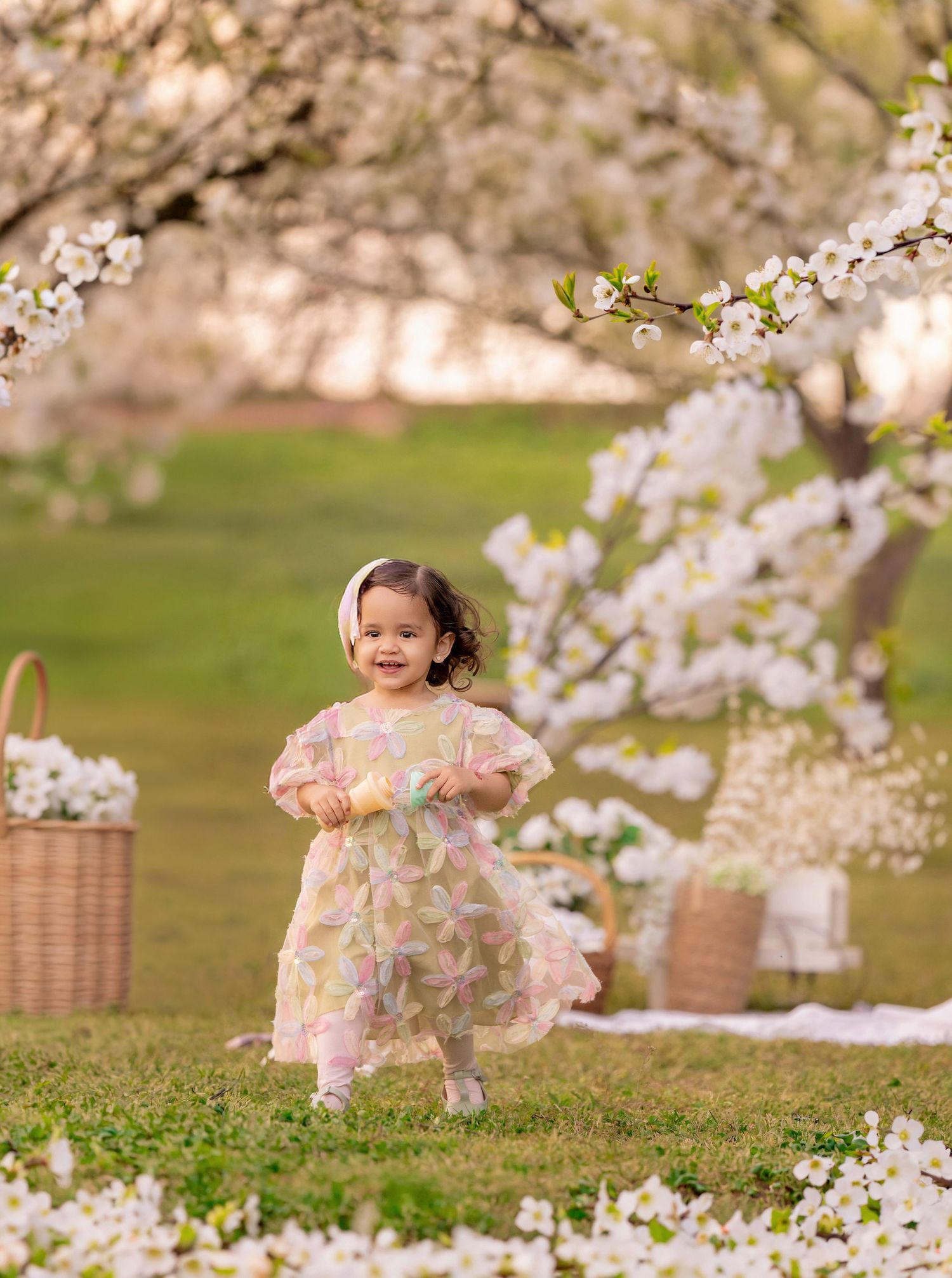 Toddler girl in floral dress running under cherry blossom trees in spring garden with wicker baskets.