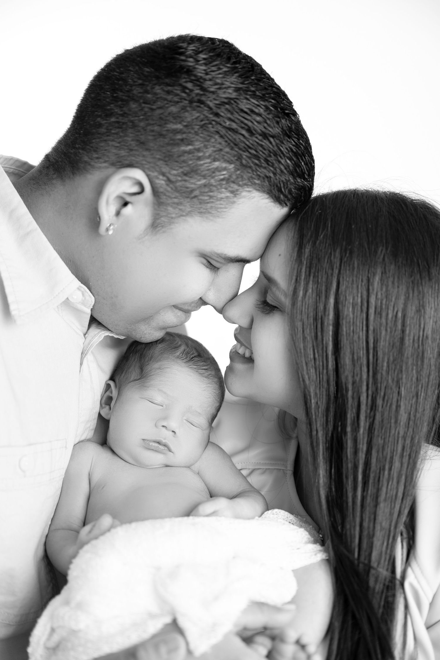 Black and white photo of new parents touching foreheads while lovingly cradling their sleeping newborn baby.