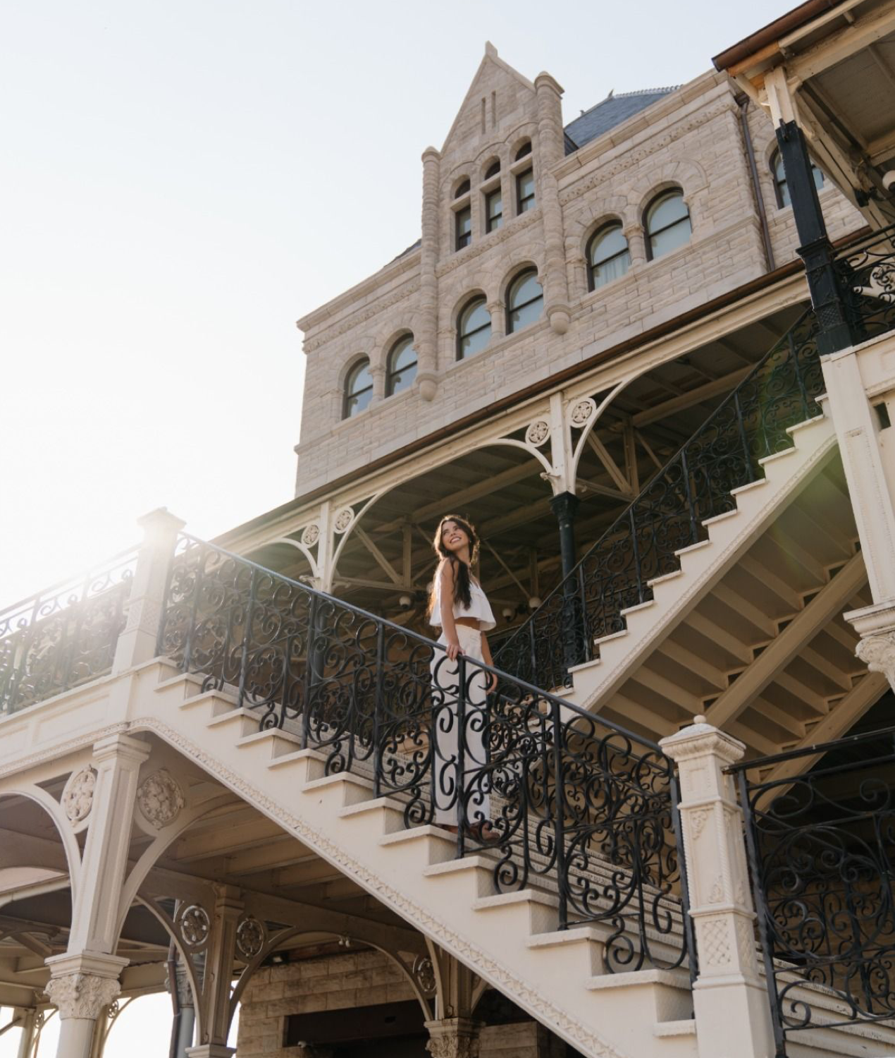 Woman standing on ornate iron staircase of historic Victorian building with arched windows in warm sunlight.