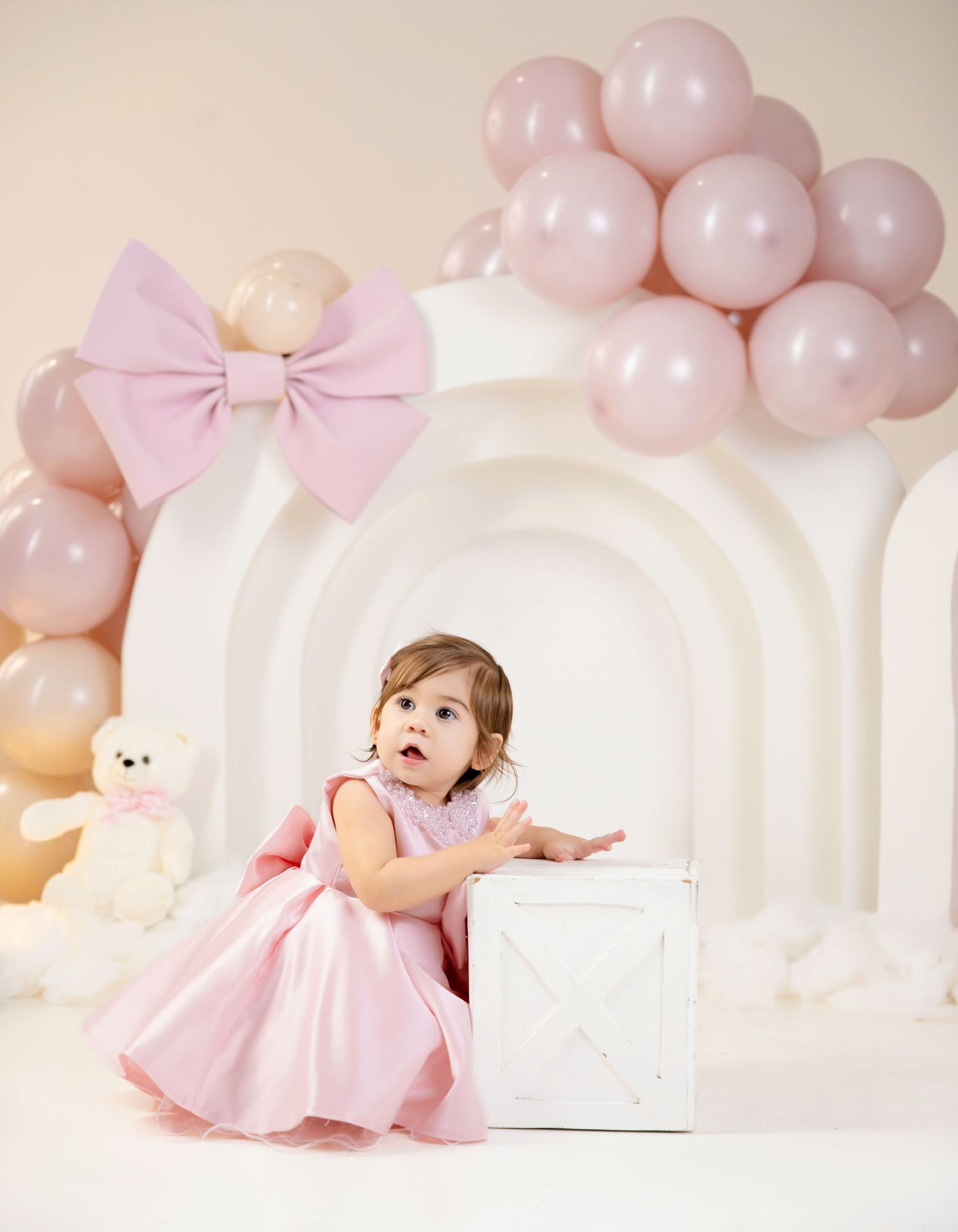 Toddler girl in pink dress sits by white box with pink balloon arch and teddy bear in birthday photo setup.