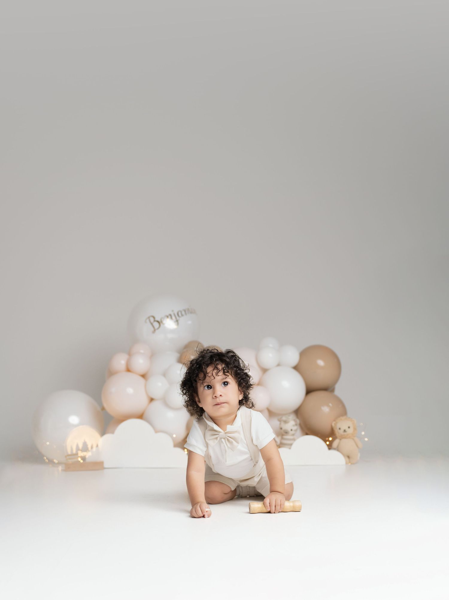 Toddler boy crawling on white floor surrounded by neutral beige and white balloon garland at a first birthday photo shoot.
