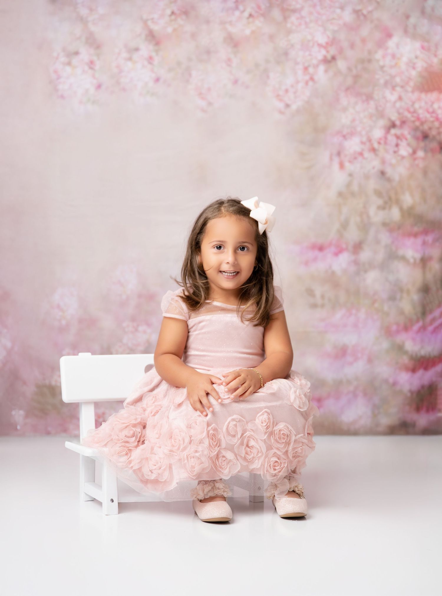 Young girl in pink floral dress sits on white bench before a soft pink floral backdrop.