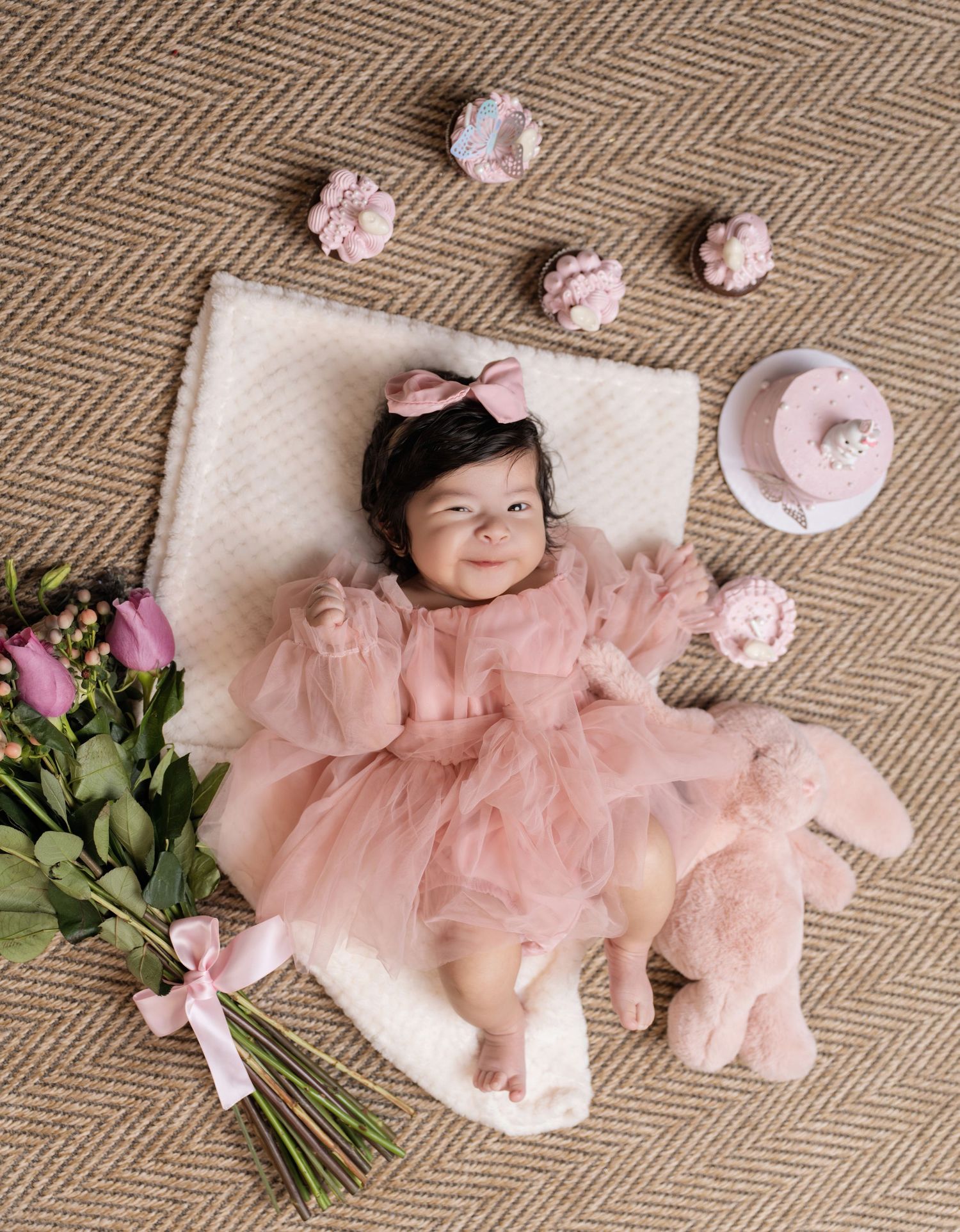 Baby girl in pink tulle dress smiles on white fur rug surrounded by flowers, cake, and a stuffed bunny.