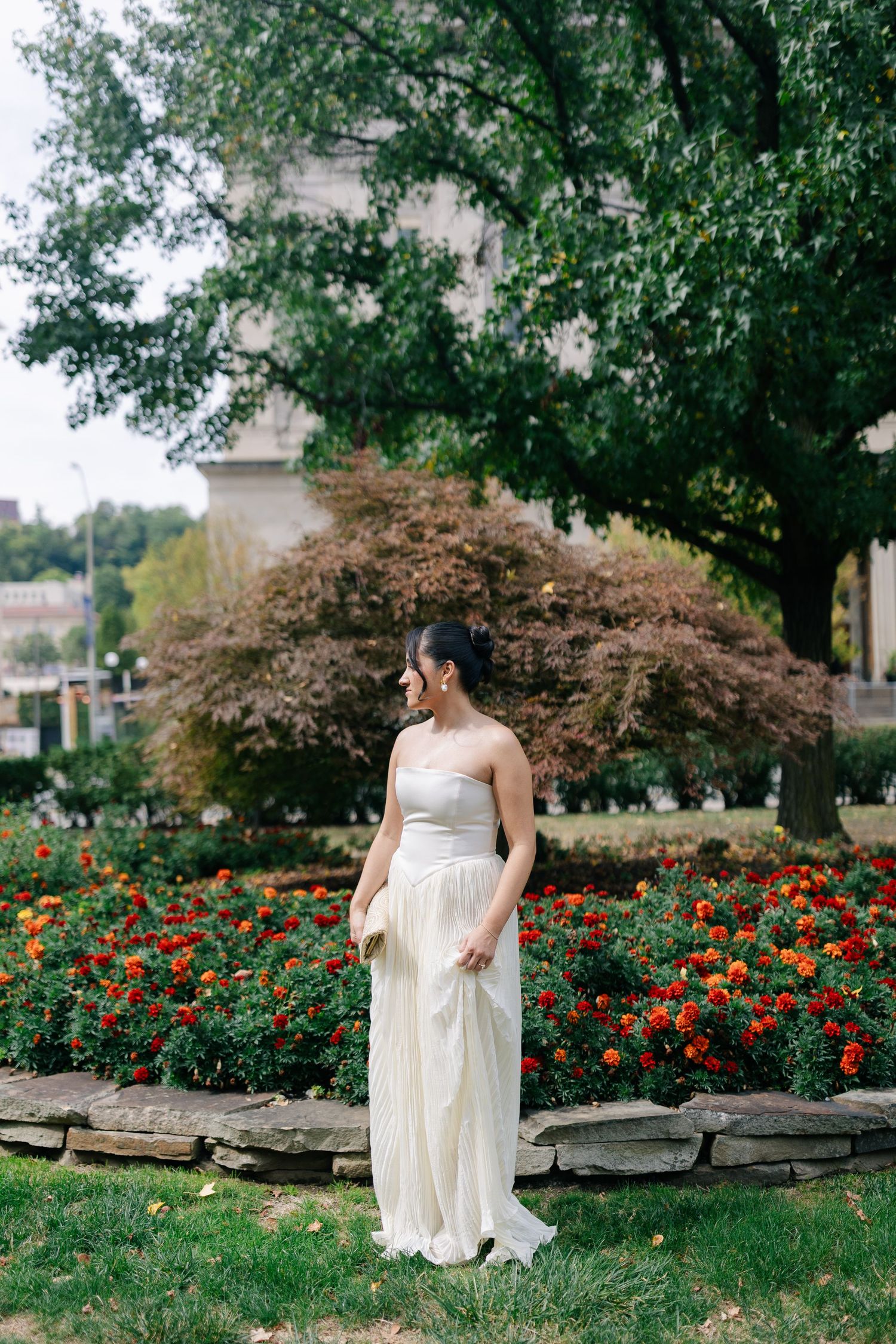 A bride in an elegant white dress stands in a garden with blooming red flowers and lush trees in the background.