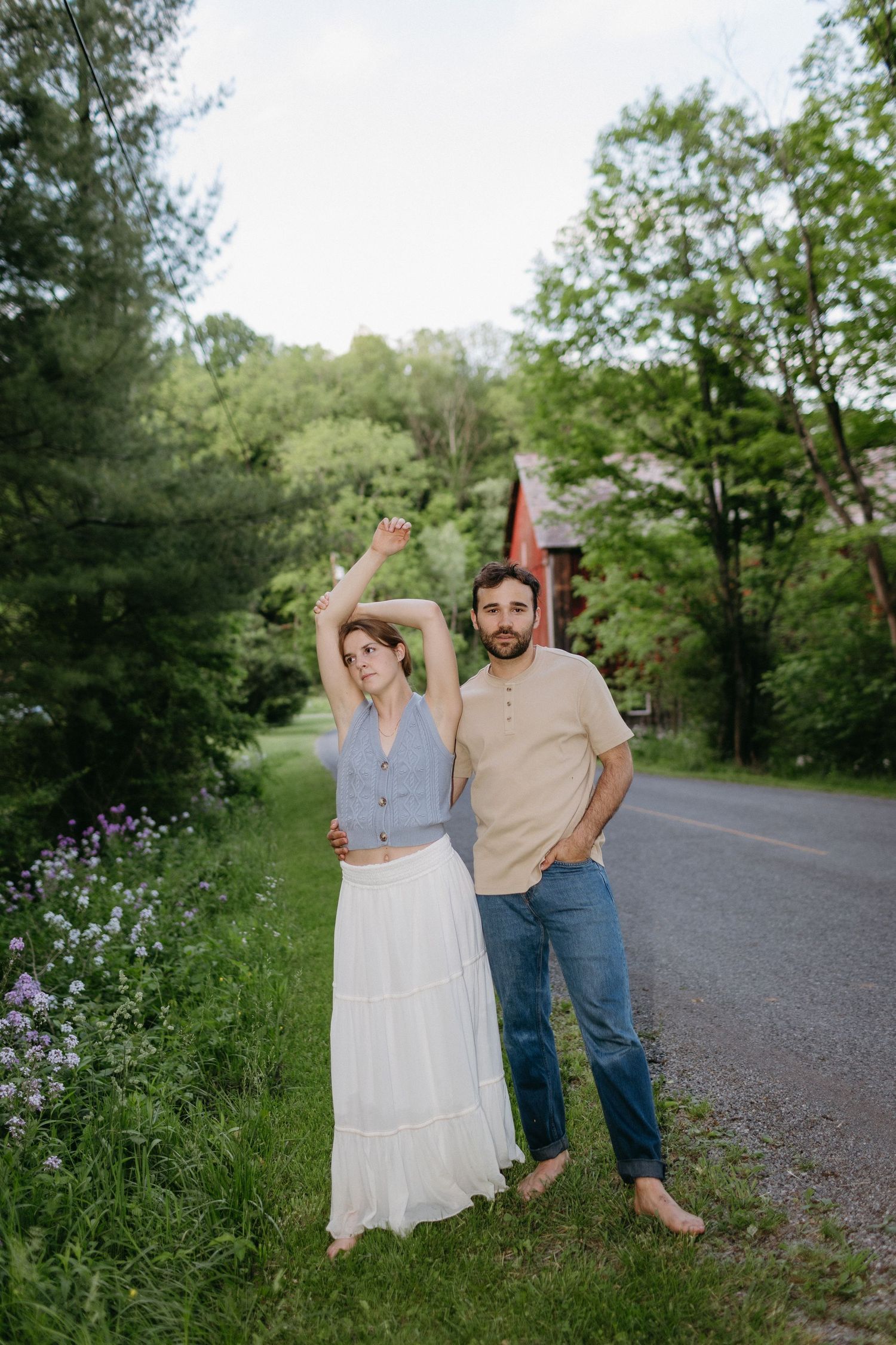 Couple posing casually on rural road with greenery and flowers.