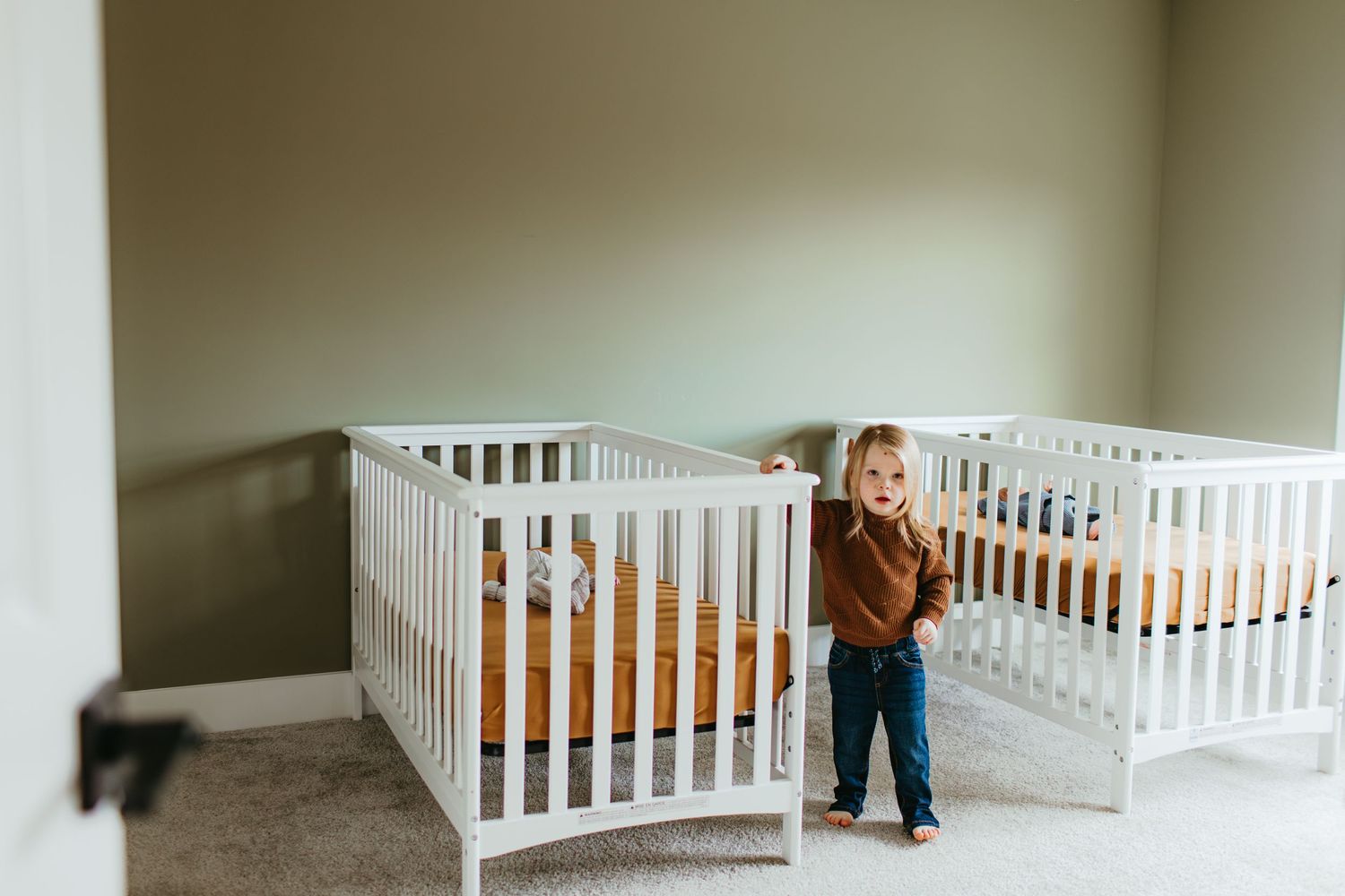 A nursery room with two white cribs and natural lighting coming through the window.