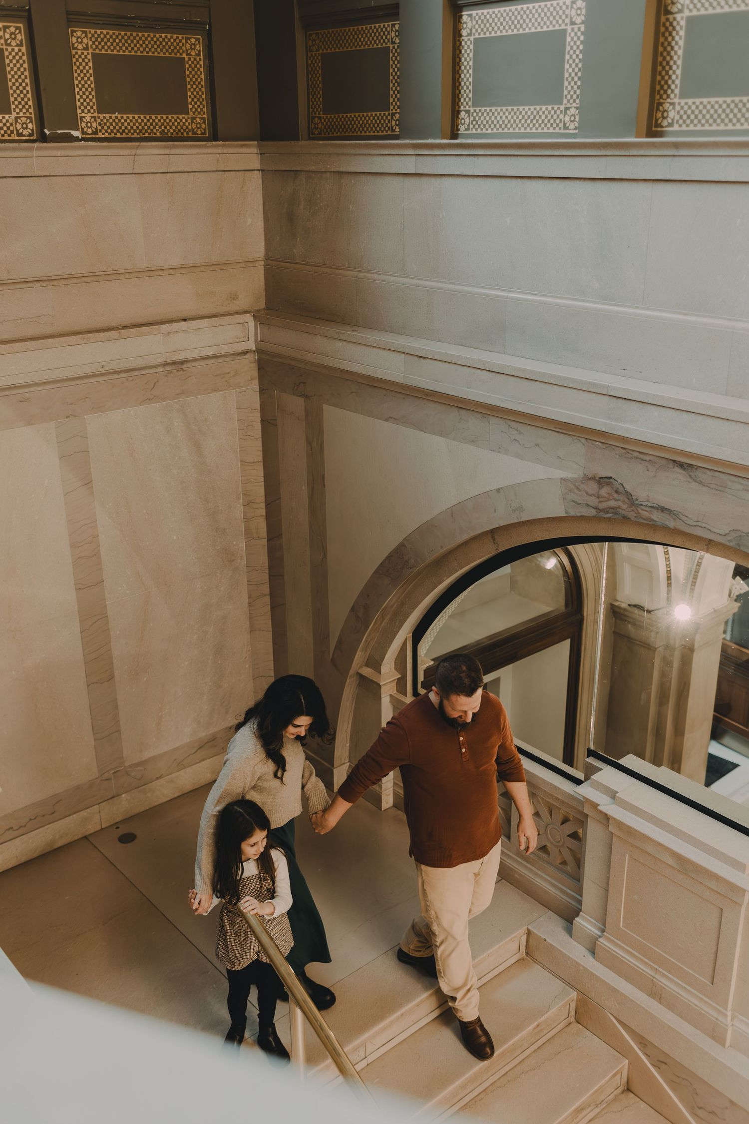 A couple walks down an elegant staircase with decorative wall molding and an arched window.