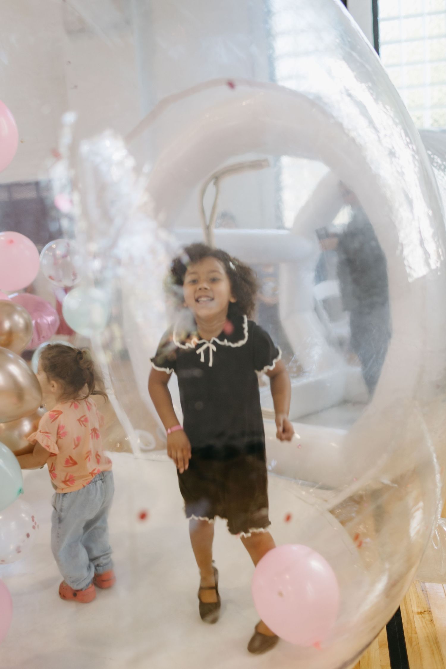 Children playing inside a transparent bubble with pink balloons at a party.