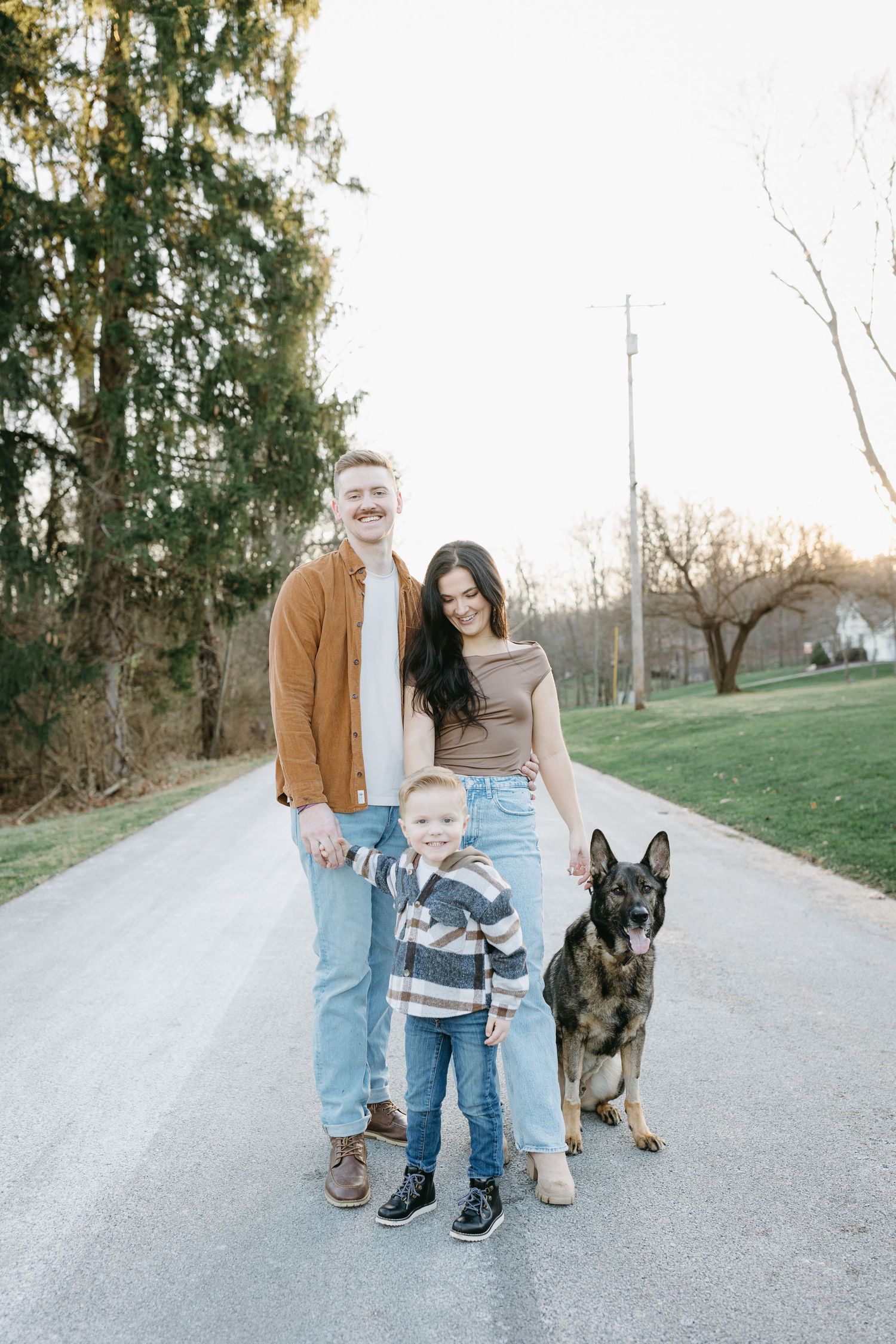 Family and their German Shepherd dog pose together on a rural road during golden hour.