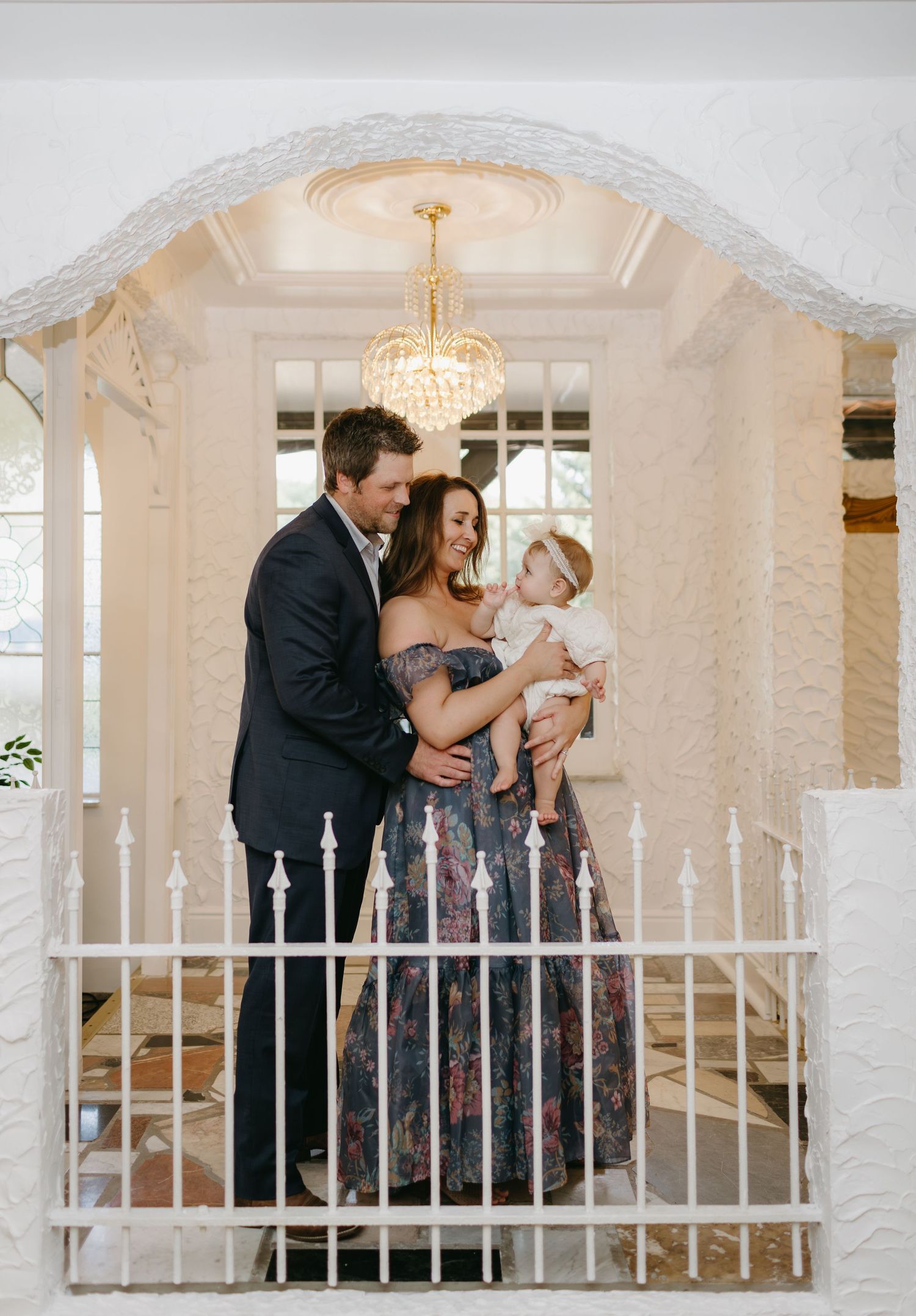 Intimate family moment under elegant chandelier and white archway with decorative railing.