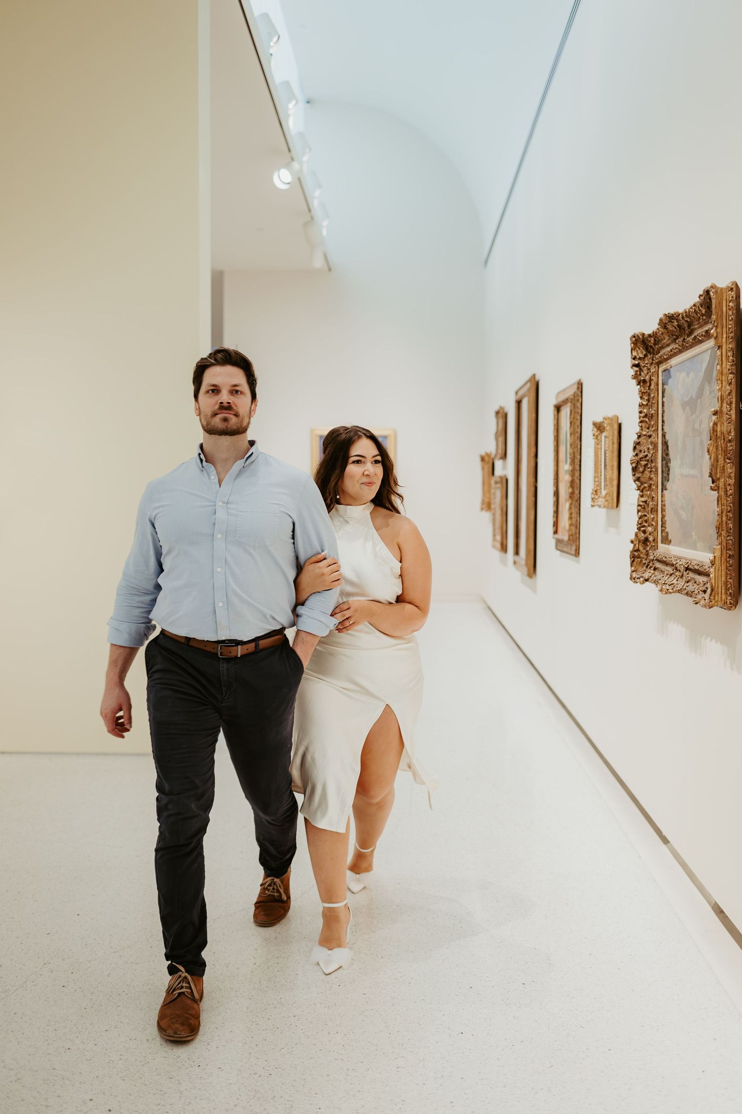 Couple exploring an art gallery with ornate gold frames hanging on white walls.