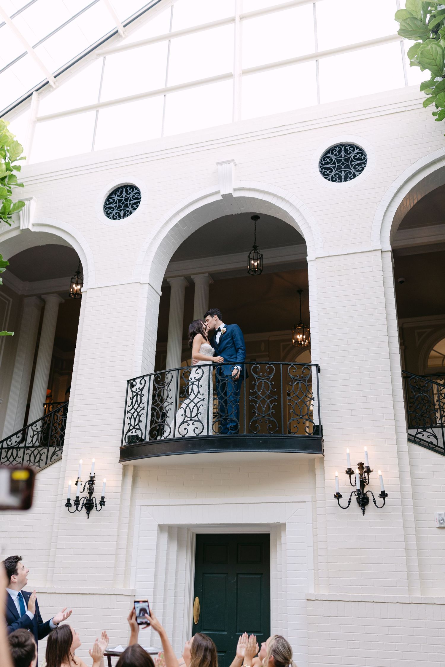 Wedding celebration at elegant white venue with couple posing on ornate balcony as guests take photos below.