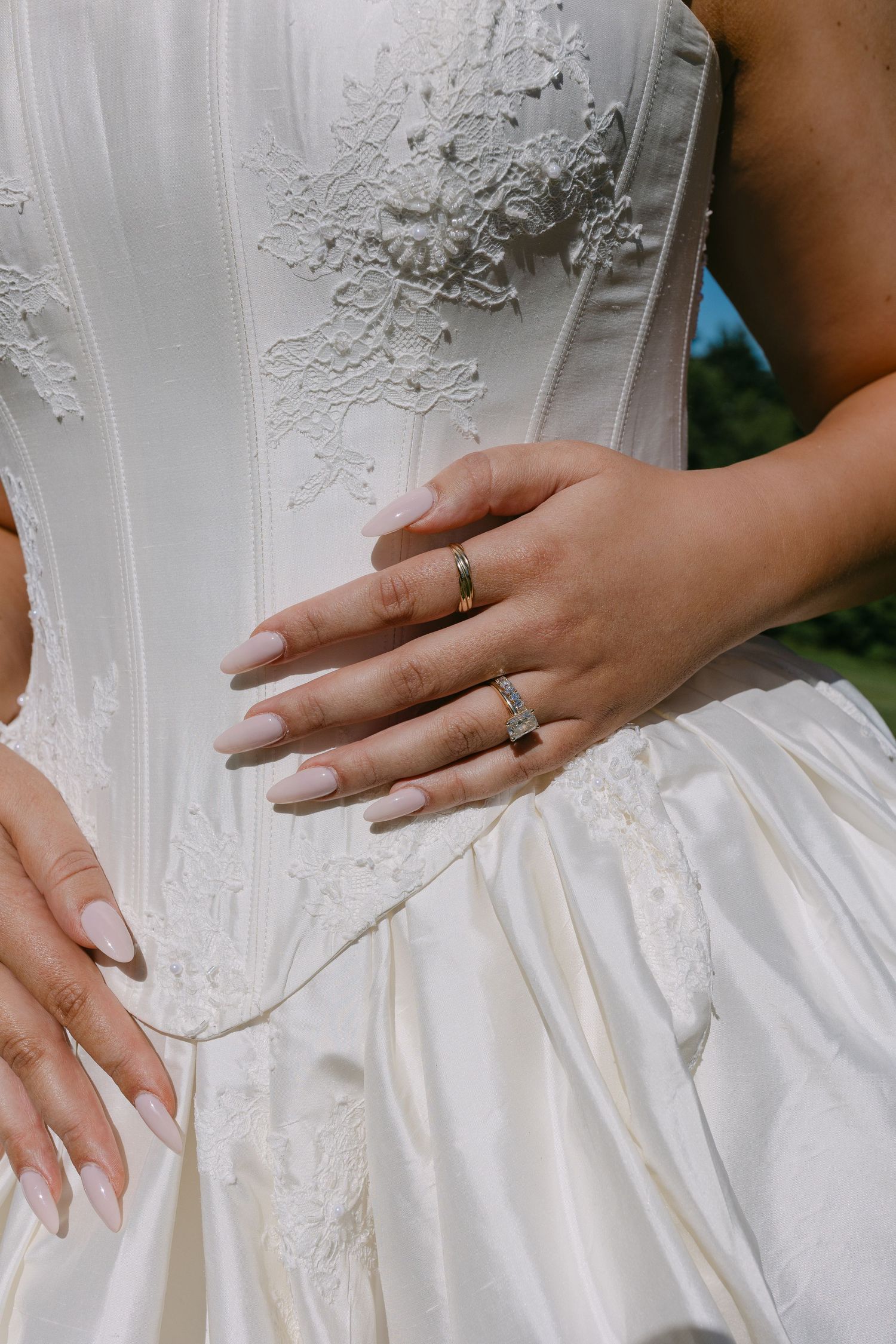 Bride's hands with elegant nude nails and gold wedding rings resting on a white lace corset wedding gown.