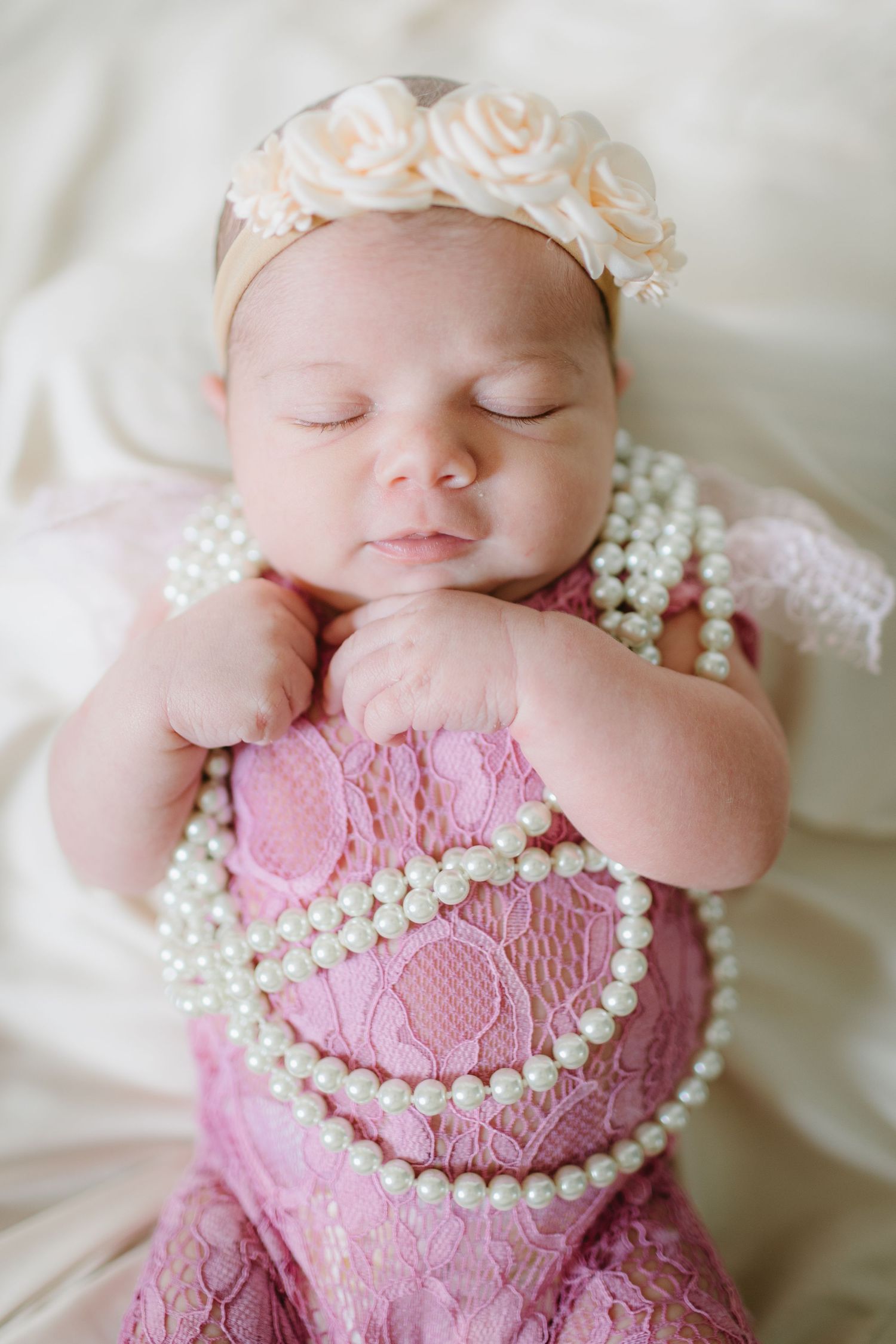 Sleeping newborn wearing pearl necklace and pink lace outfit with floral headband.