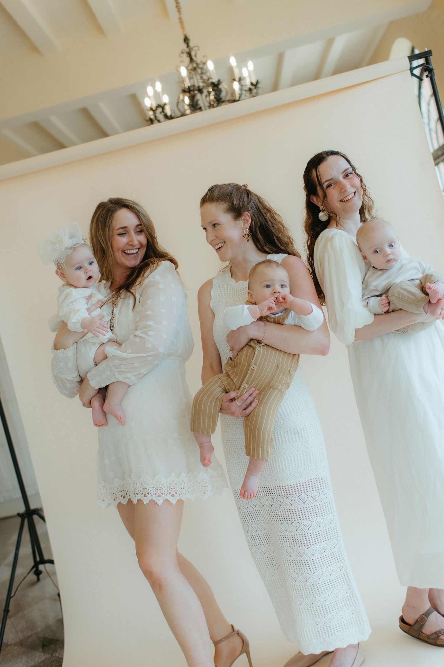 Three women in white dresses hold infants in bright studio setting.