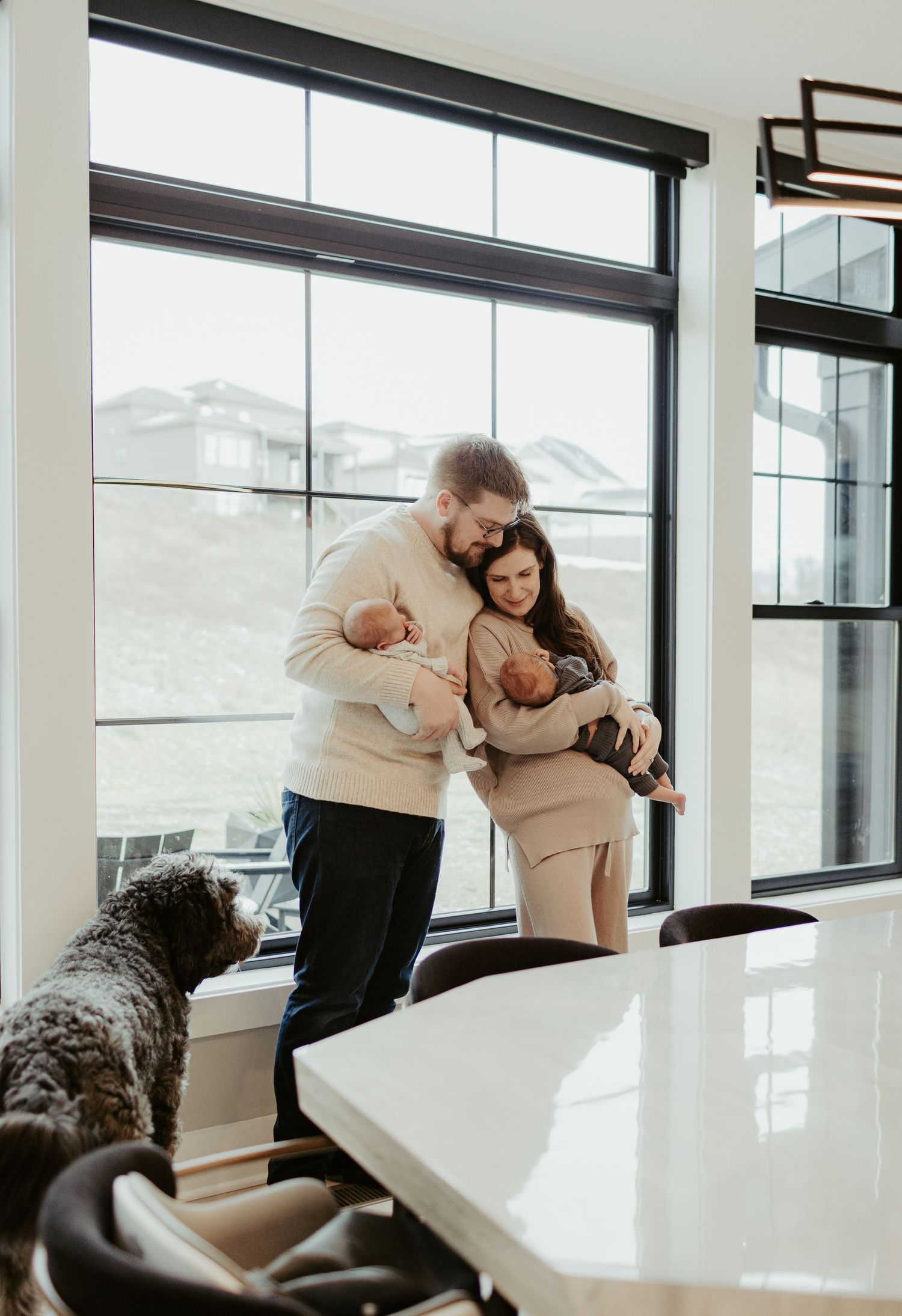 A couple in cream sweaters gazes out a large window at a snowy mountain landscape with their dog nearby.