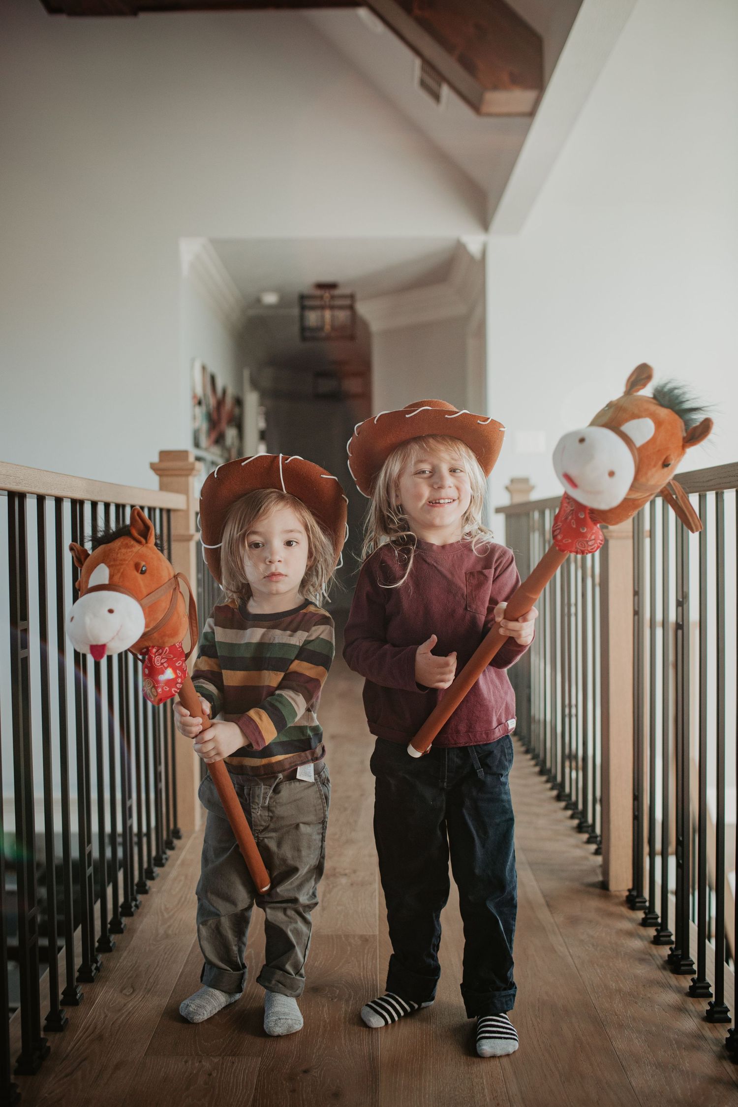 Two small children in cowboy hats holding stick horses in a hallway.