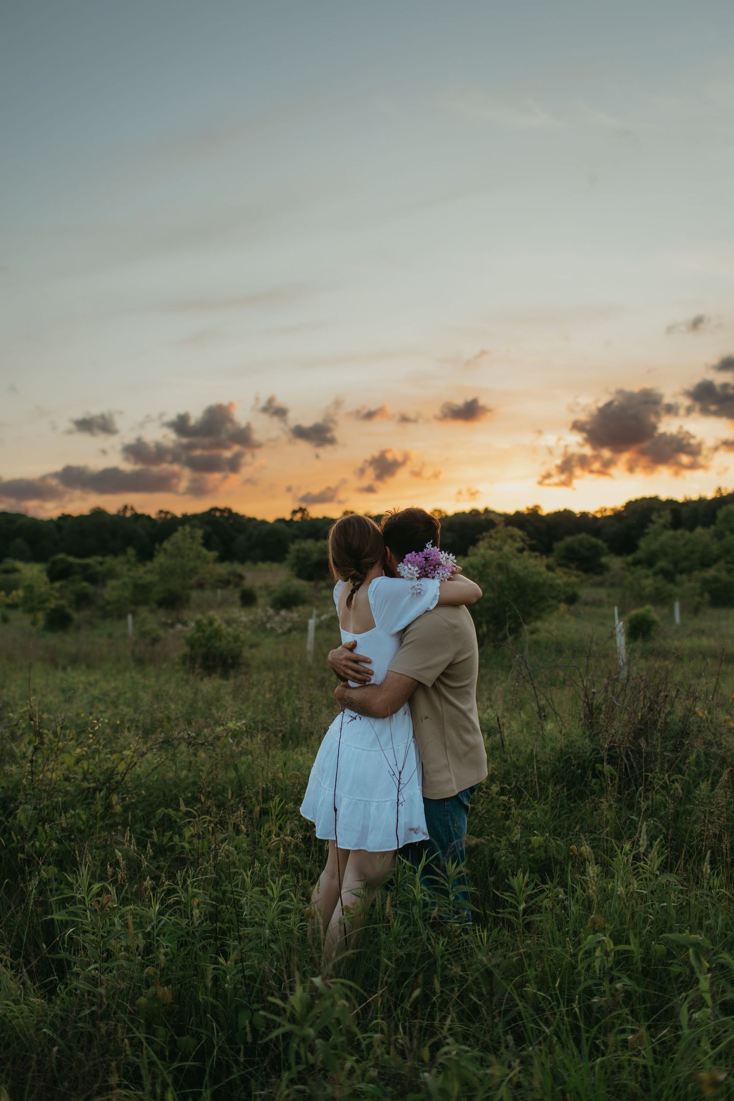 Couple embracing in a field at sunset with golden light filtering through trees.