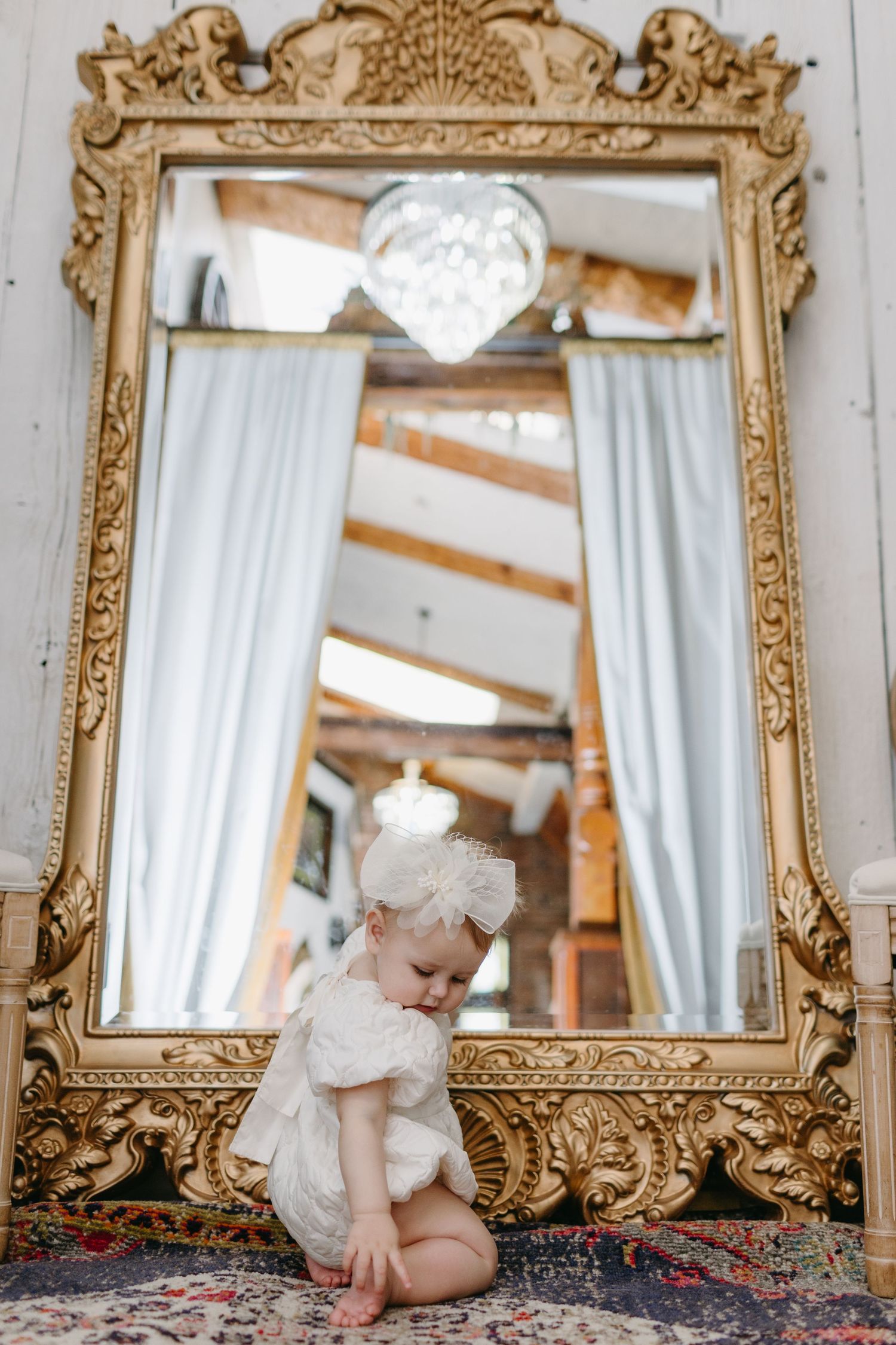 Baby in white dress sits beneath ornate gold mirror with decorative frame.