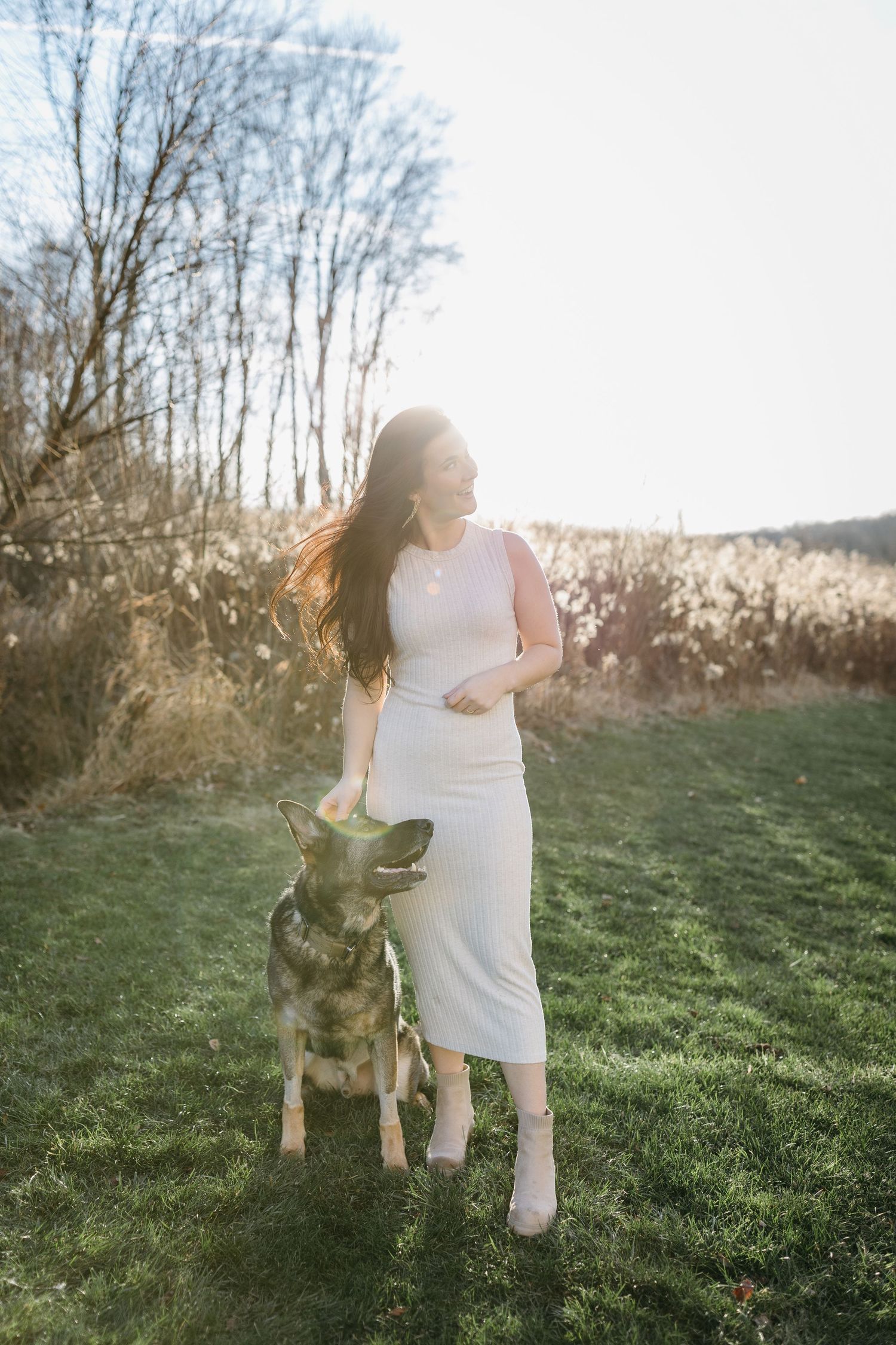 Woman in white dress poses with German Shepherd dog in sunlit field.