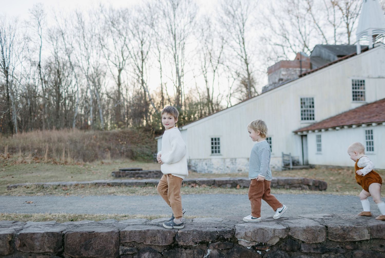 Children walking along a stone wall outside a white farmhouse at dusk.