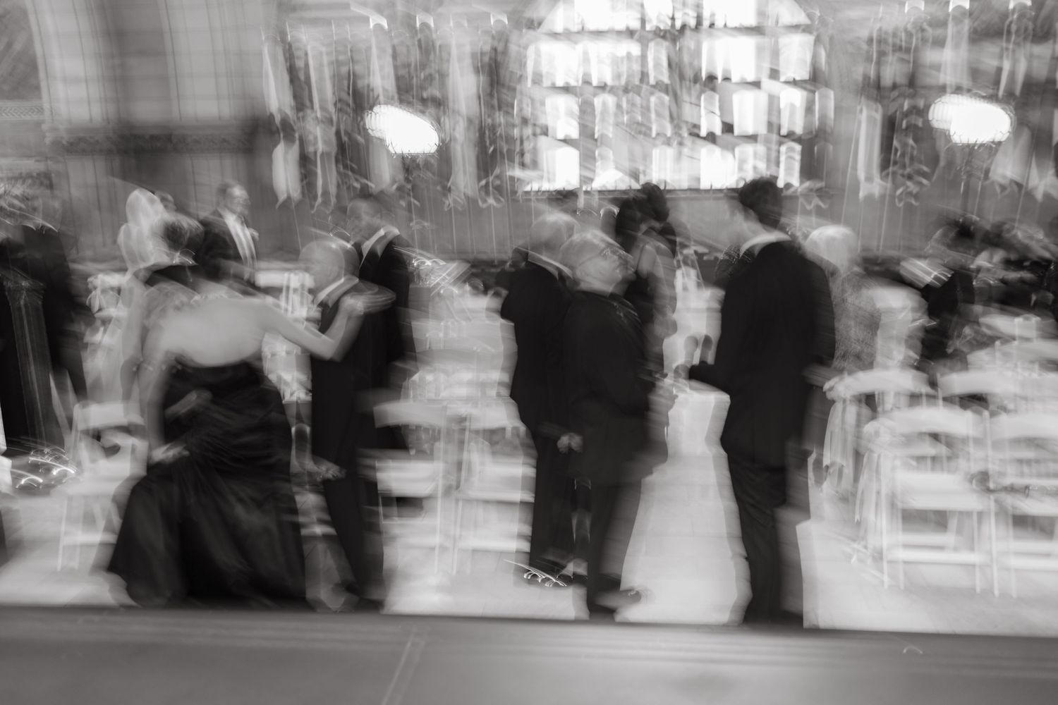 A blurred black and white image of people gathering in an elegant indoor space, creating a dreamy vintage atmosphere.