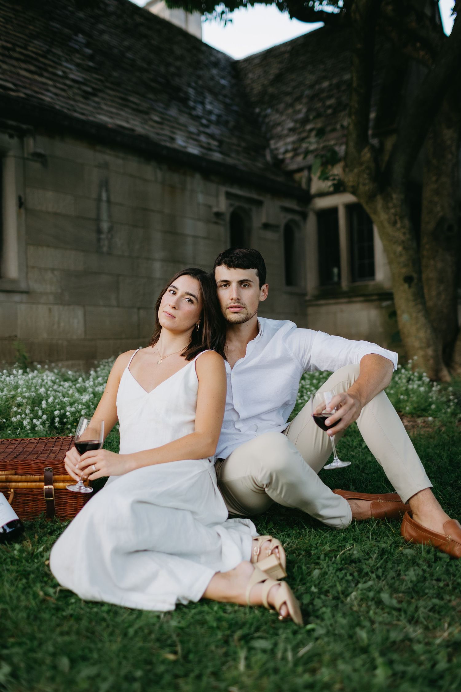 Romantic outdoor picnic scene with couple in white outfits lounging on grass near stone building.