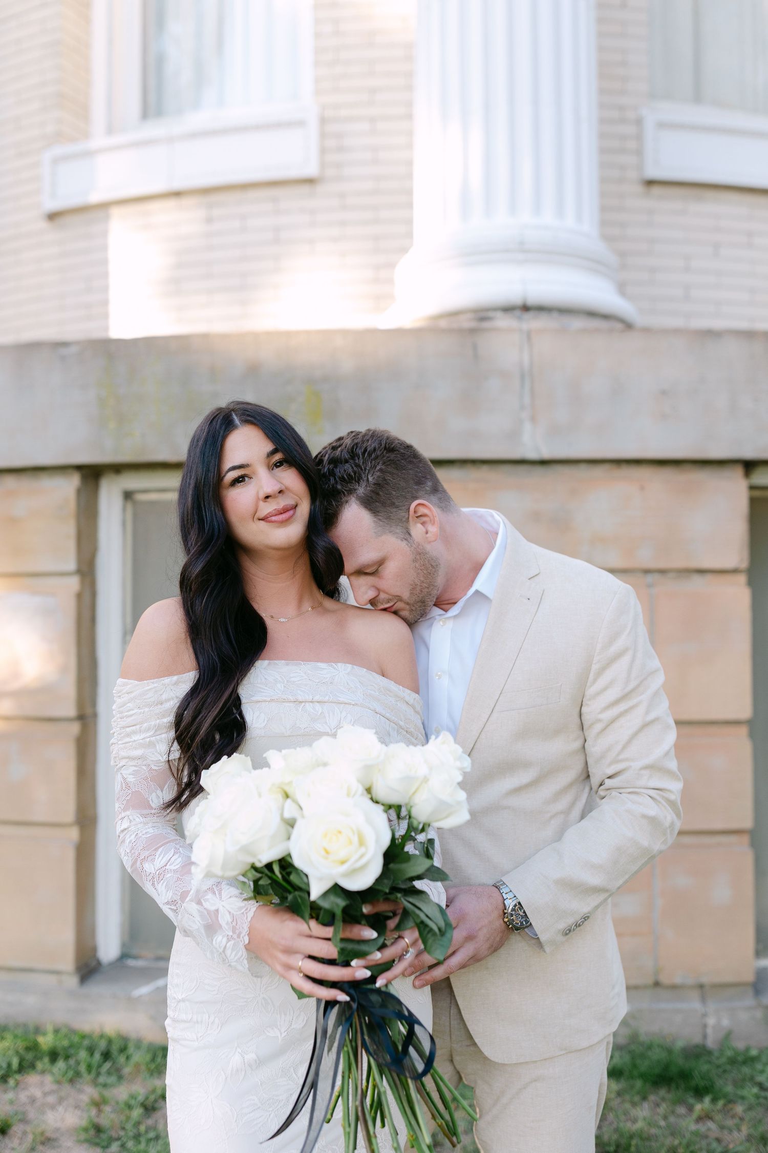 A romantic wedding moment captured outdoors with white roses and elegant off-shoulder lace dress against a classical building facade.