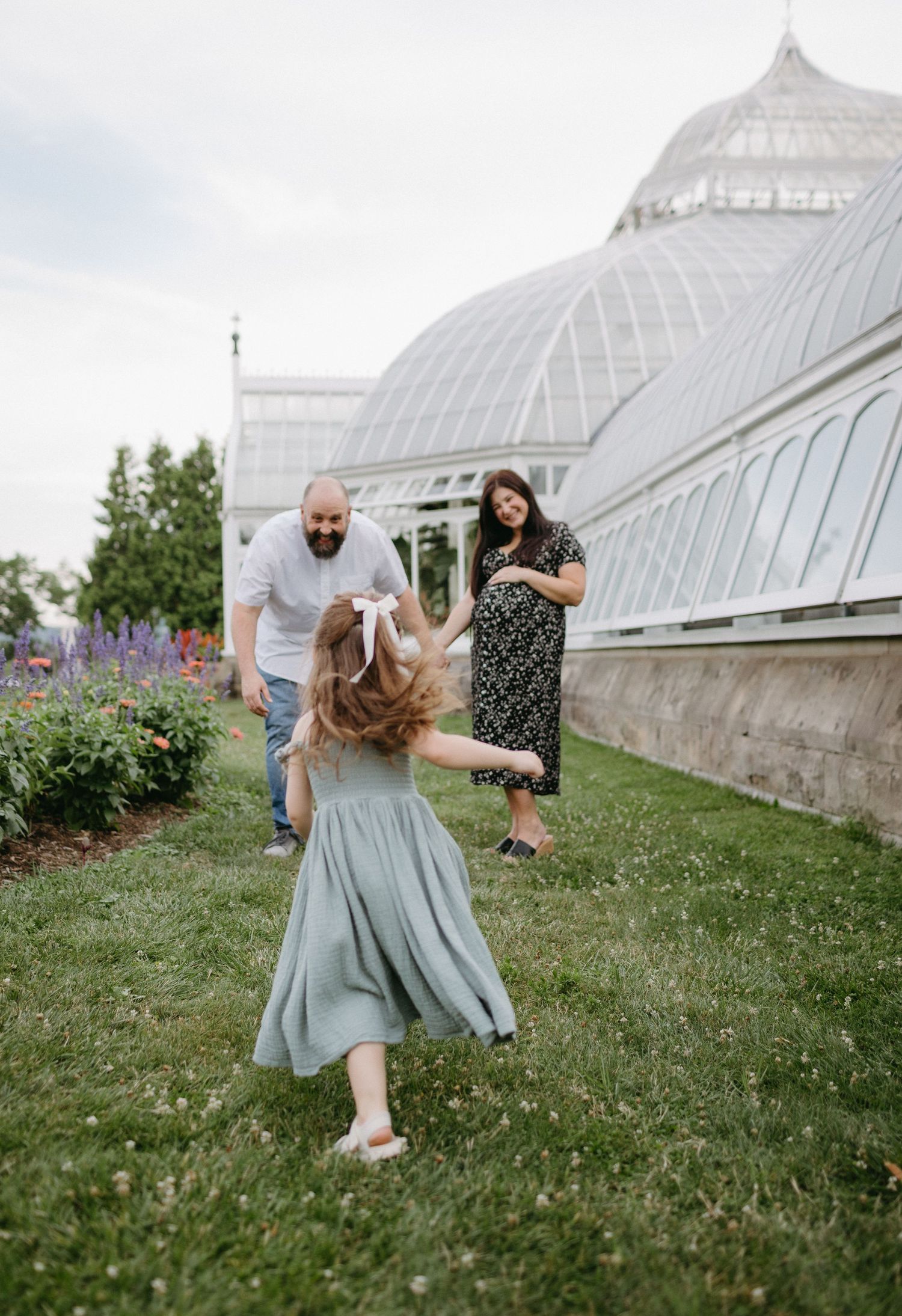 Playful family scene on grass with Victorian-style greenhouse in background.