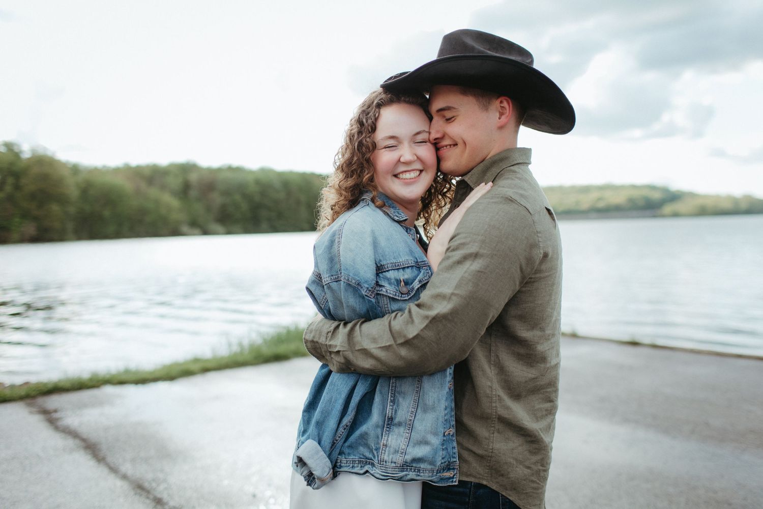Couple in casual wear embracing by lakeside, one wearing cowboy hat.