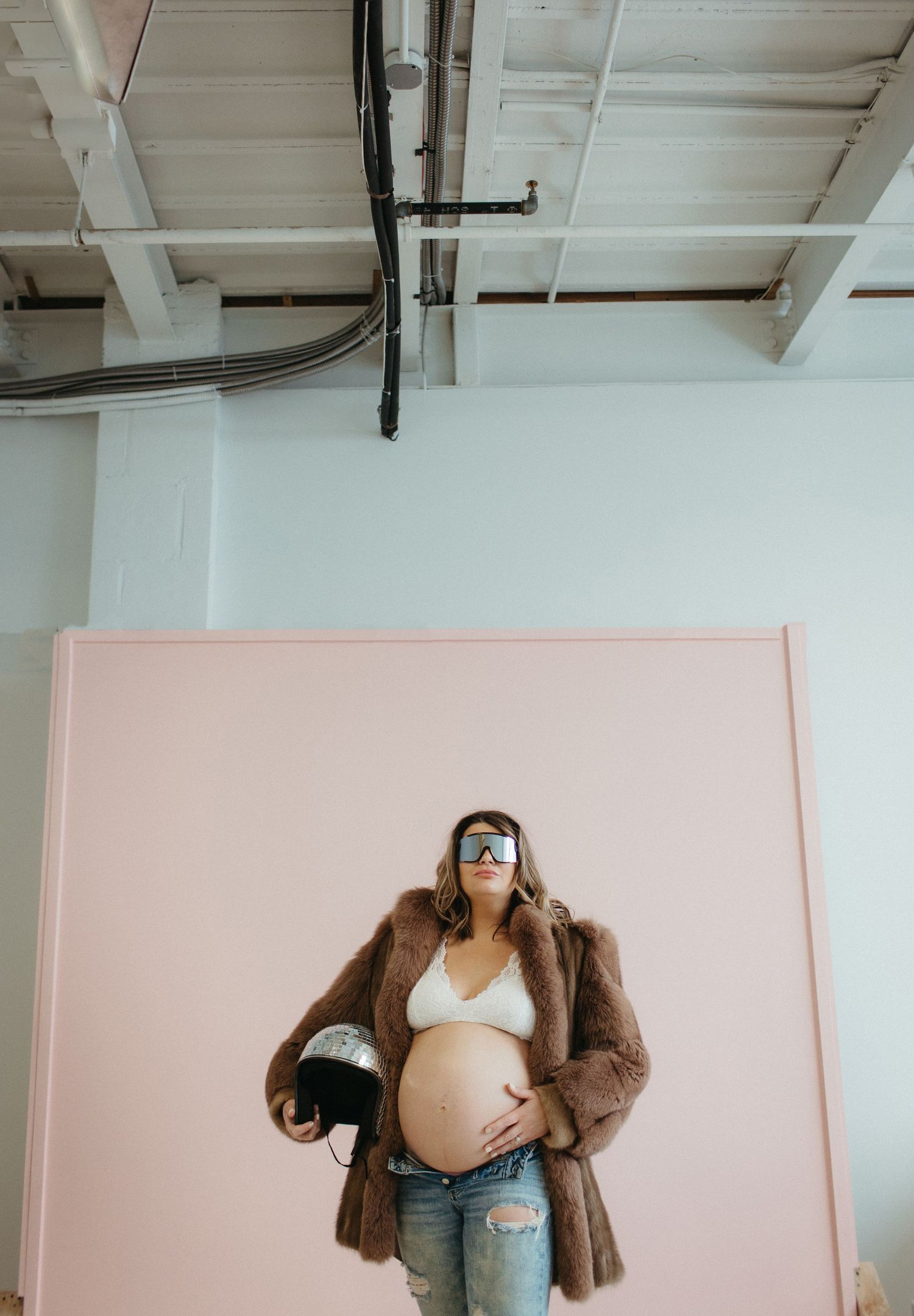 Stylish maternity photo with pregnant woman in fur coat against pink backdrop.