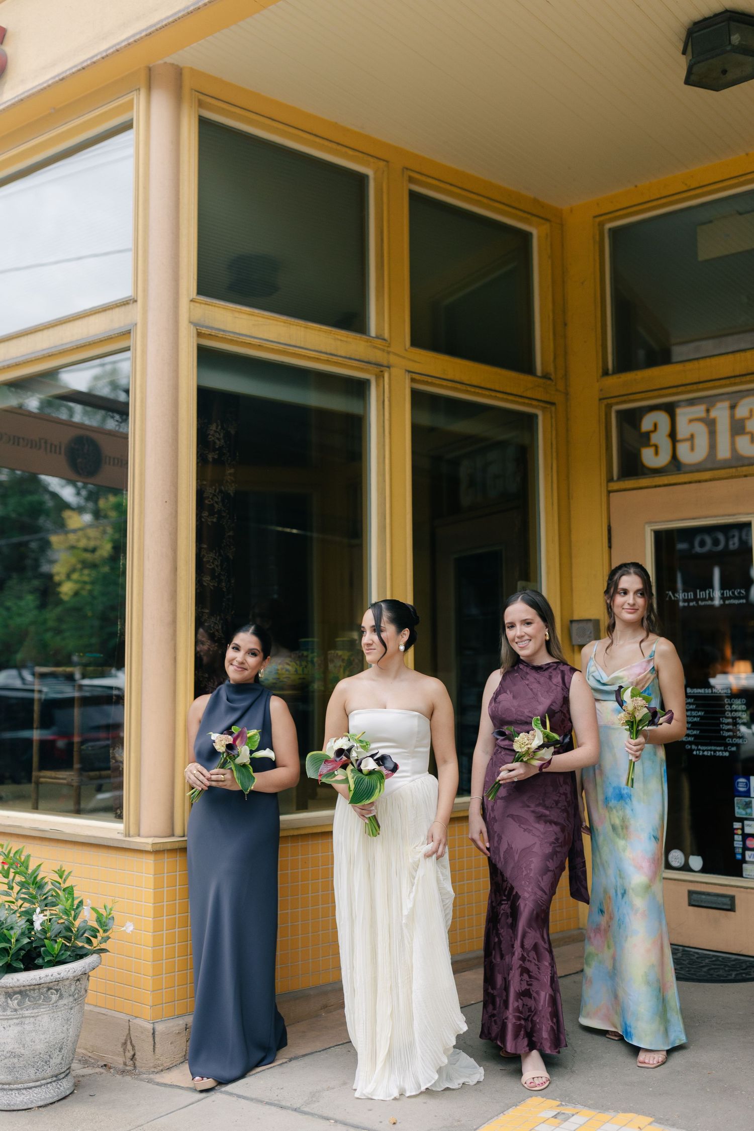 Wedding party in formal attire stands outside yellow building with glass windows.