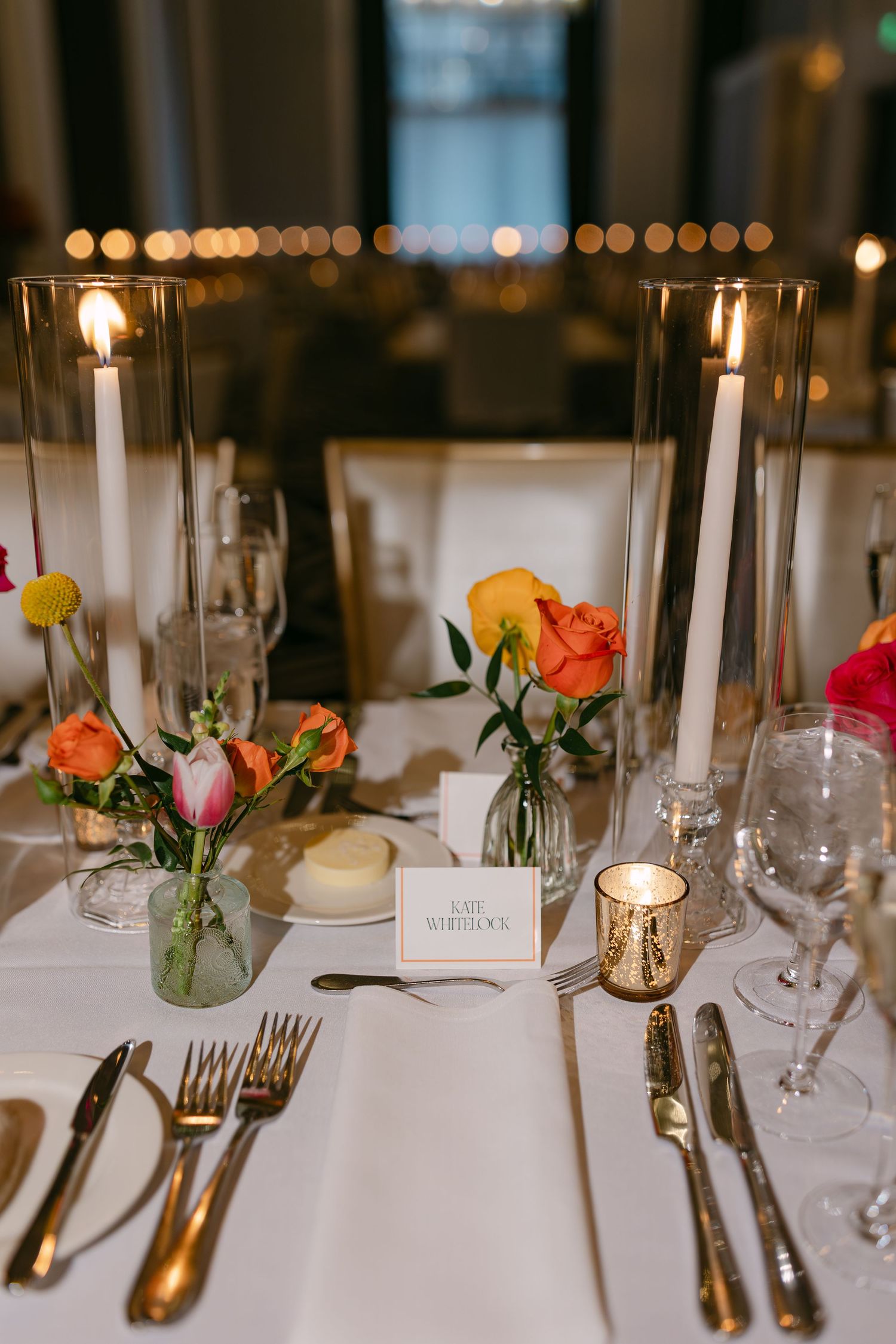 Elegant wedding table setting with taper candles, small rose bouquets in glass vases, and gold flatware on white linens.