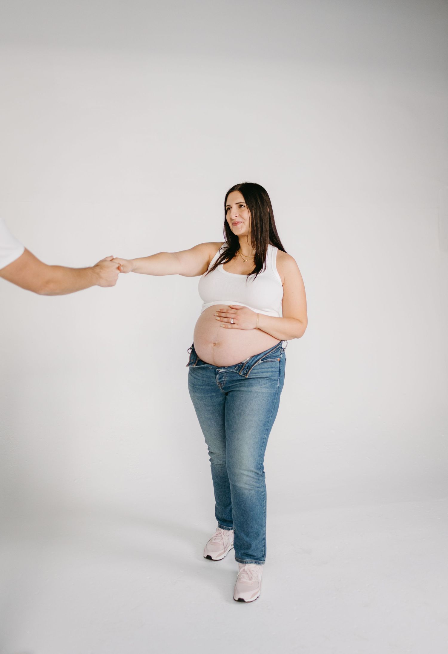 Expectant mother in casual attire holding hands with someone off-camera against white backdrop.