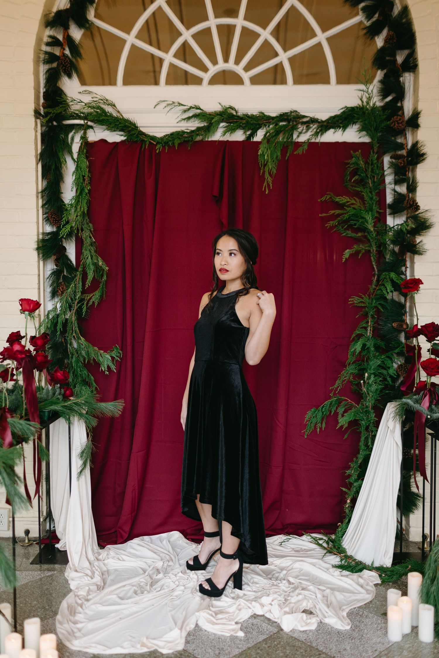 Elegant black velvet dress posed against red curtain backdrop with evergreen garland and white fabric decorations.