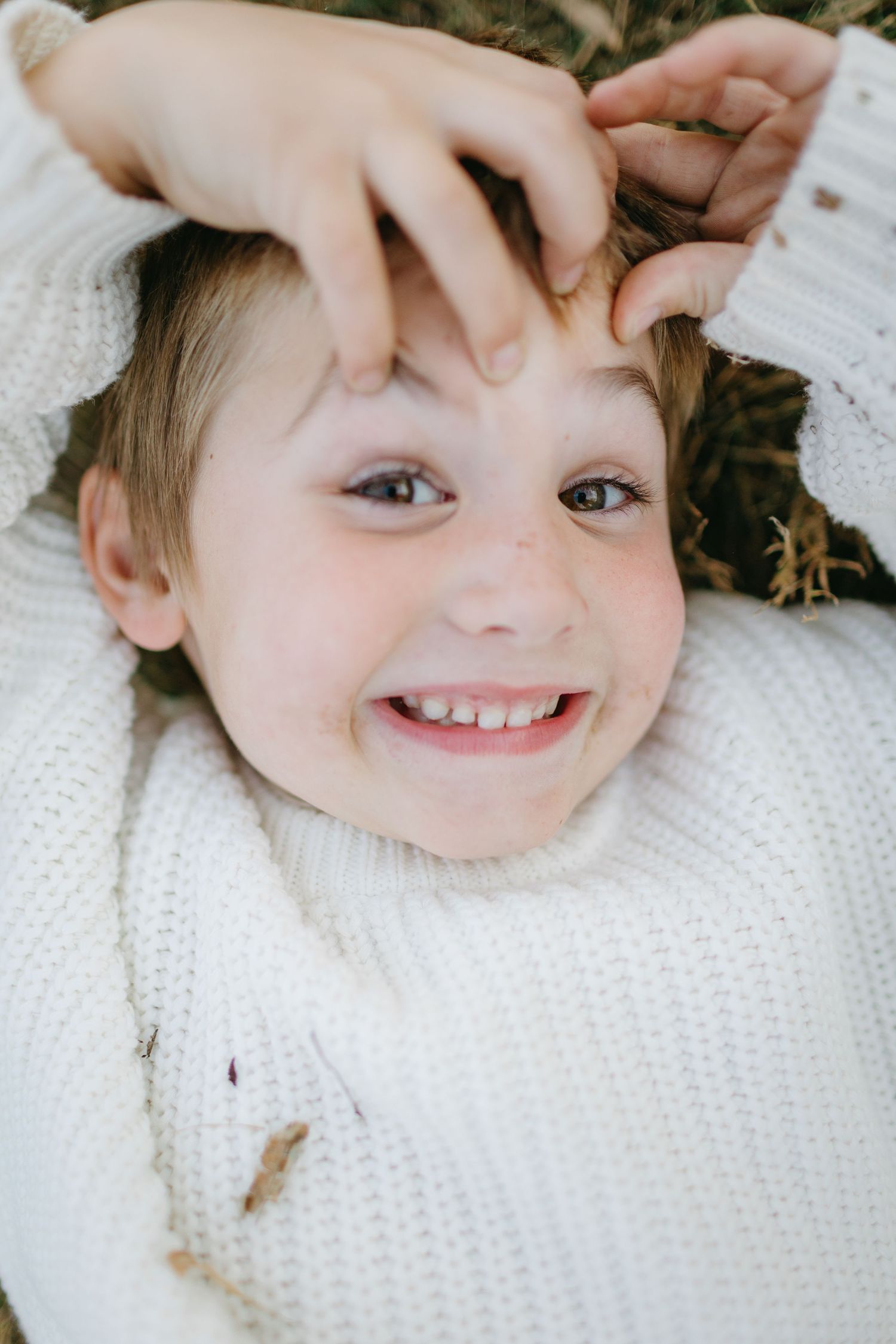 A close-up portrait of someone wearing a white sweater lying down outdoors.