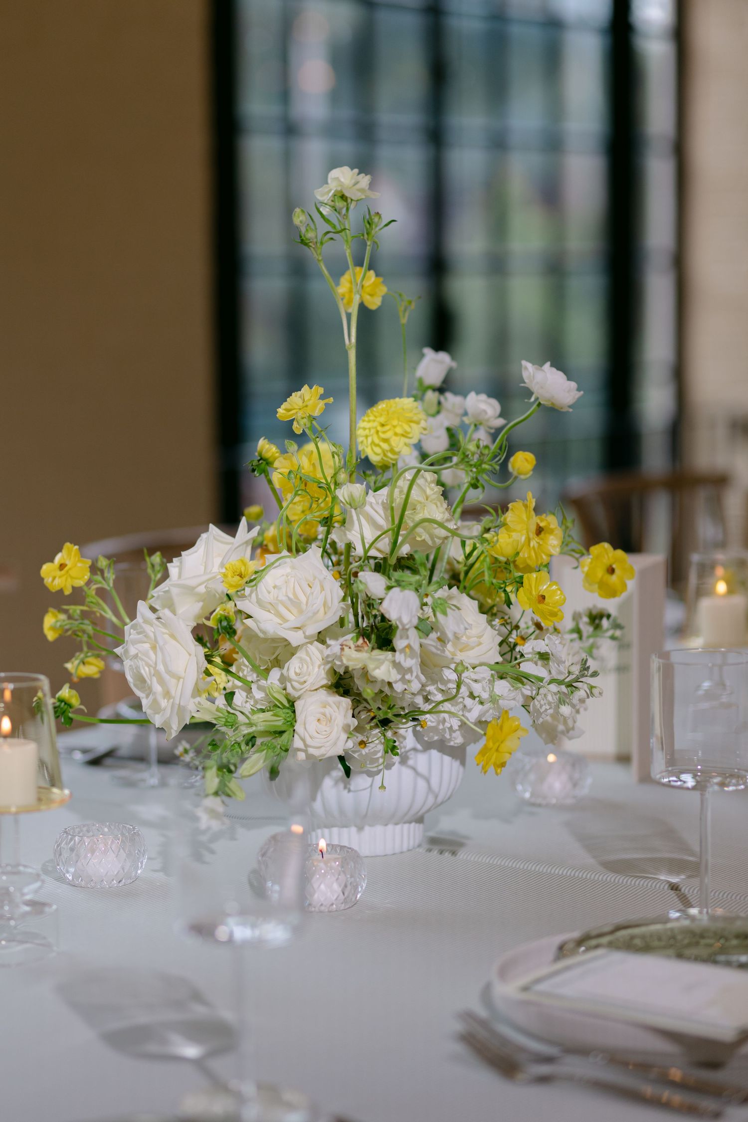 Elegant wedding centerpiece with white roses and yellow blooms in a white vase on a white tablecloth.