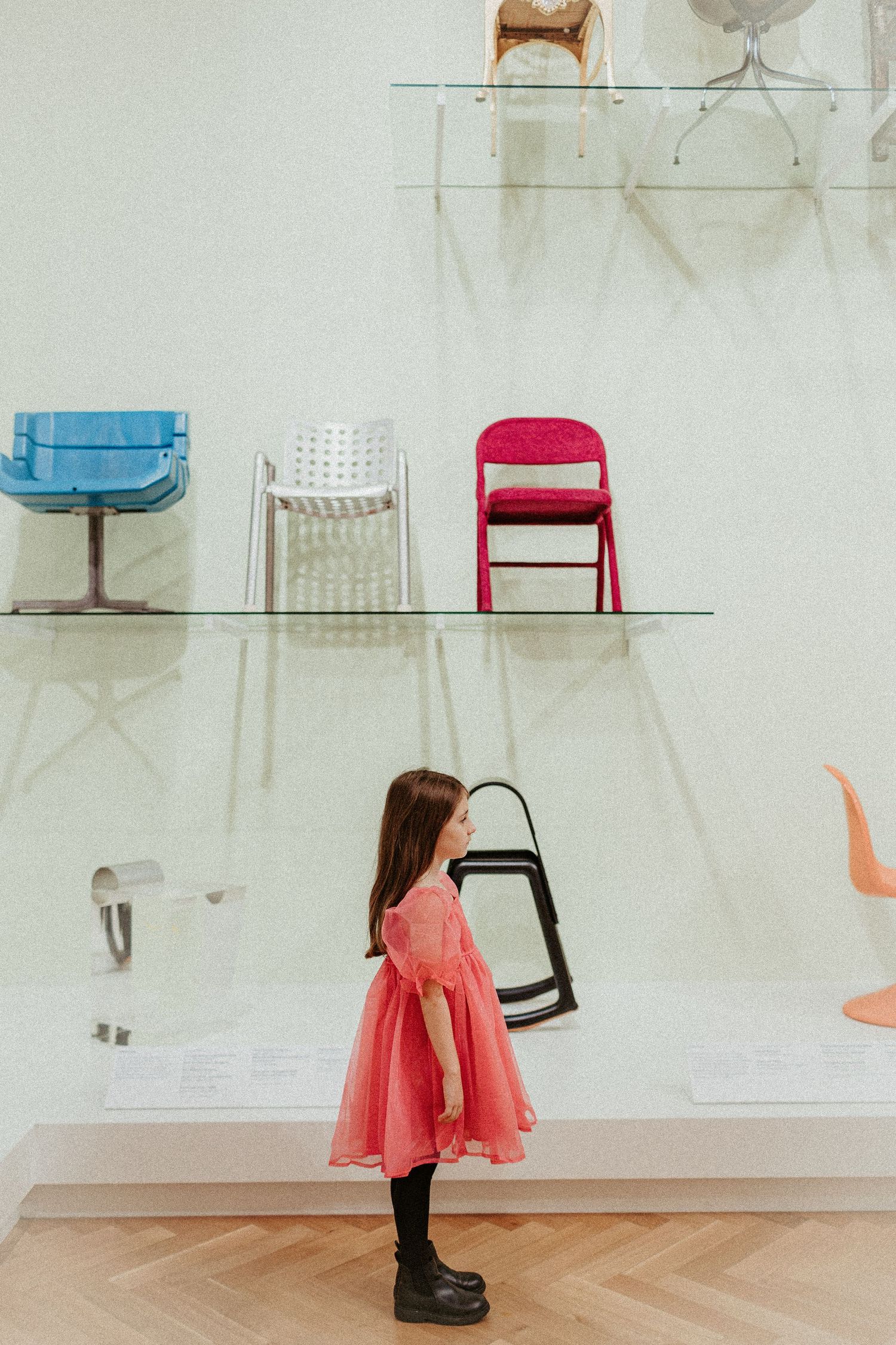 Child in coral dress looking at chairs displayed on wall shelves in modern gallery setting.