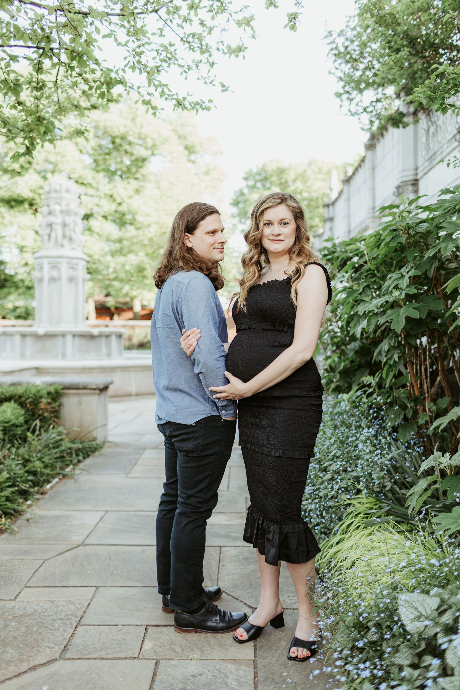 A couple poses together on a garden path, with one wearing a black dress and the other in casual blue attire.