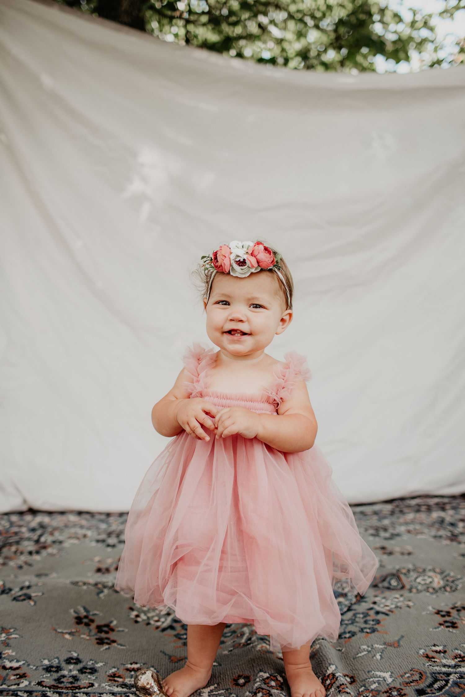 A small child in a pink tulle dress and floral headband stands against a white backdrop.