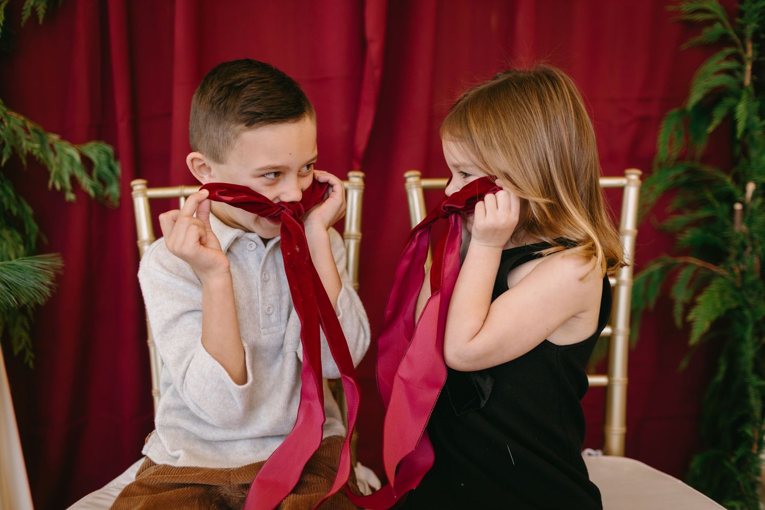 Two children playfully cover their faces with red fabric against burgundy backdrop.