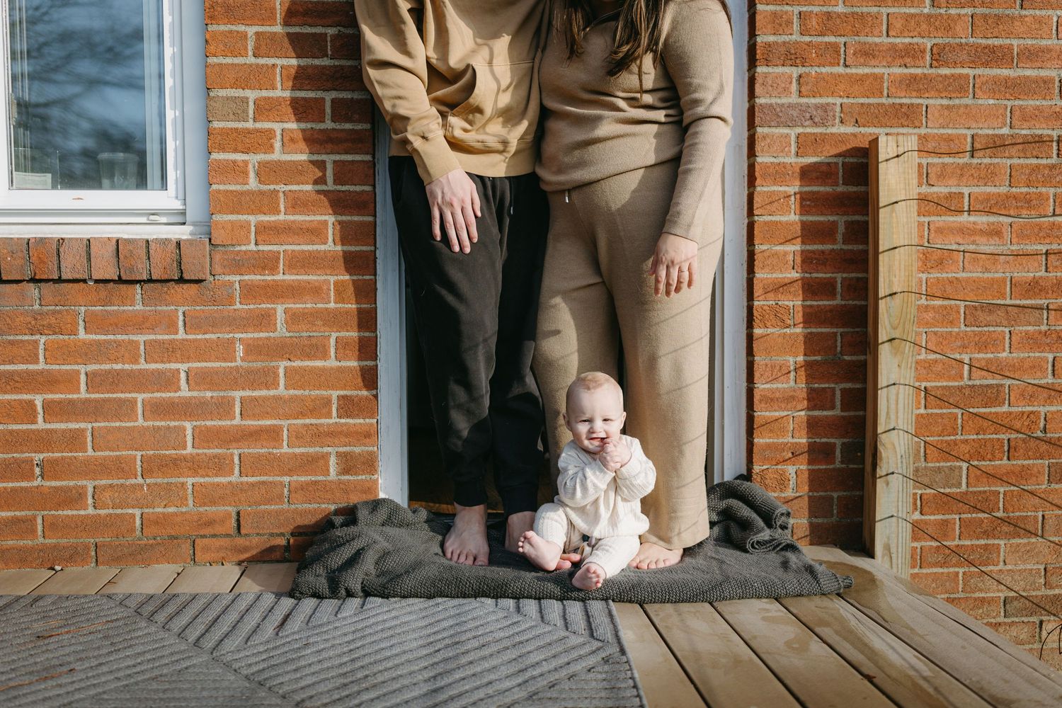 Baby sits on doorstep between adults' feet wearing light colored outfit.